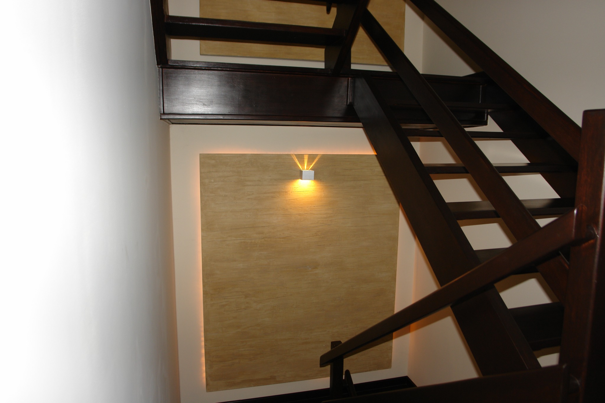 Drewniane schody w ciemnym odcieniu, oświetlone punktowym światłem na ścianie z dekoracyjną fakturą. Ujęcie z dołu, podkreślające geometrię konstrukcji.