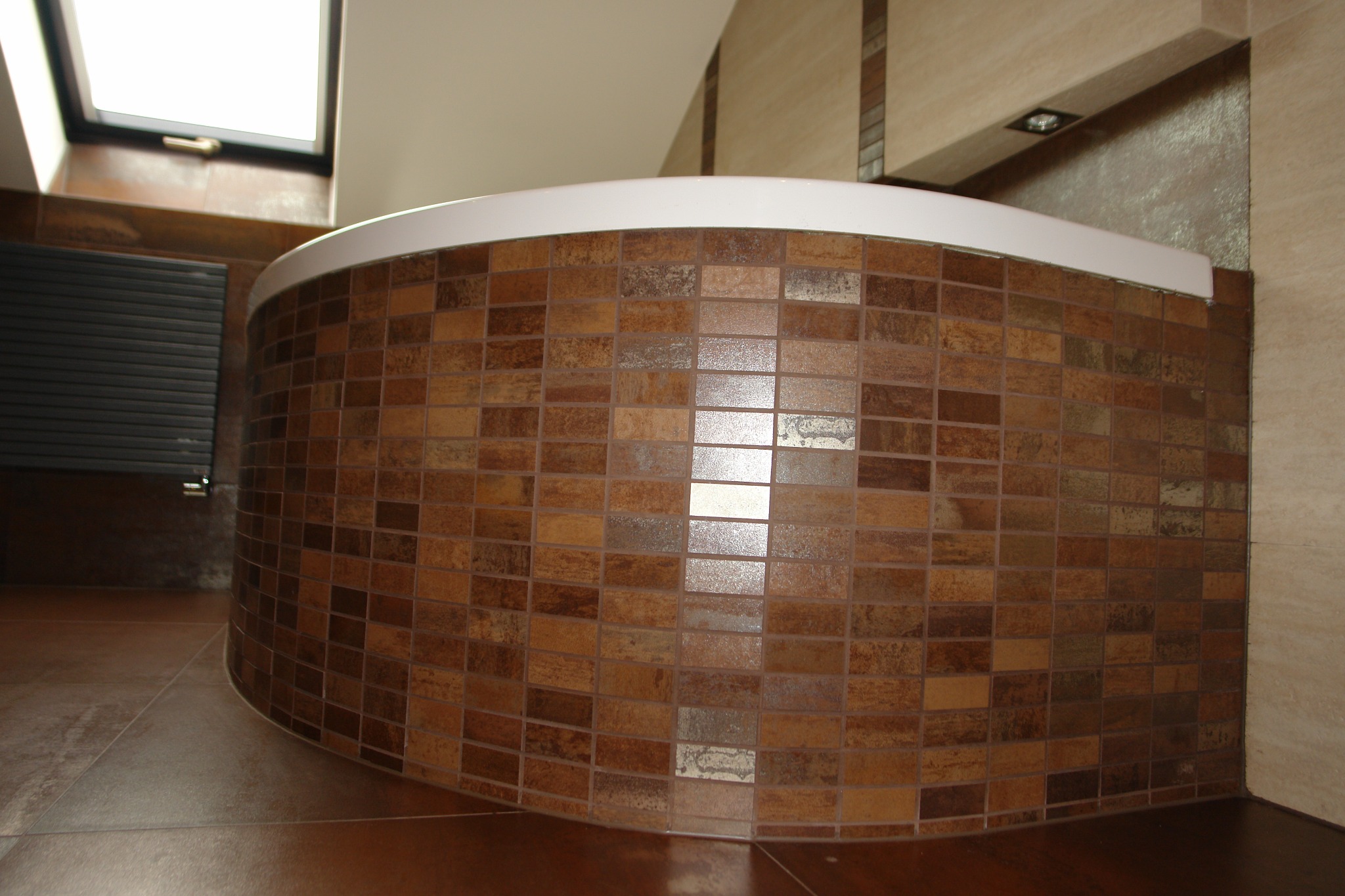 Zaokrąglona obudowa wanny wyłożona mozaiką w odcieniach brązu i beżu, z widocznym fragmentem podłogi i okna dachowego. Ciepła kolorystyka płytek.