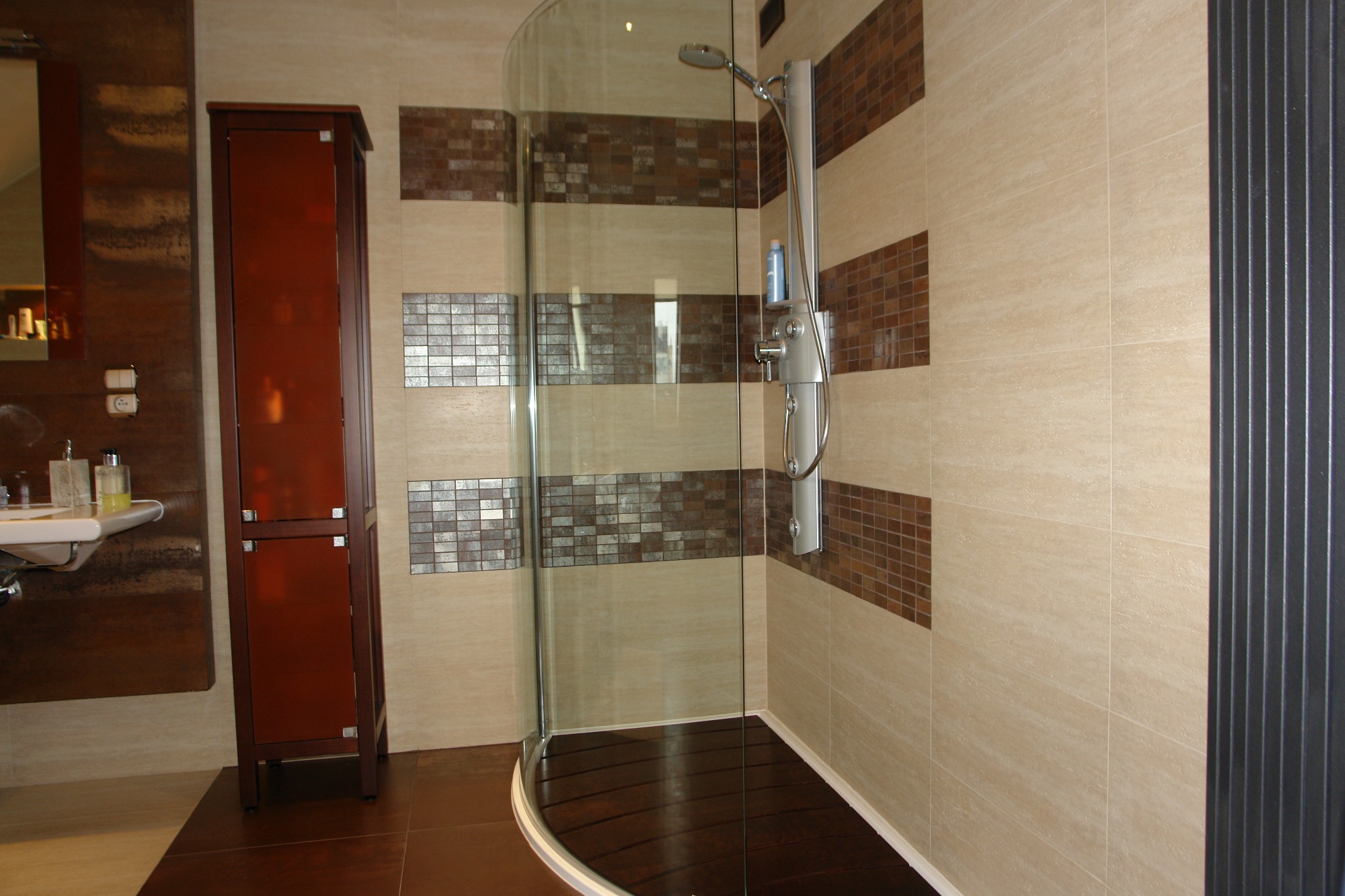 Wykończona łazienka z kabiną prysznicową z zaokrąglonym szkłem, brązowymi płytkami w pasy i szafką w kolorze ciemnego drewna. Podłoga z ciemnych paneli.