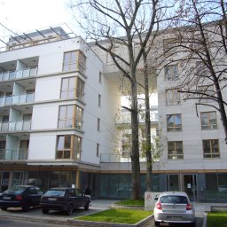 ArchiZmiana - studio architektoniczne - Nowoczesny budynek mieszkalny z balkonami i dużymi oknami, widoczny z poziomu ulicy, z zaparkowanymi samochodami i drzewami na pierwszym planie.