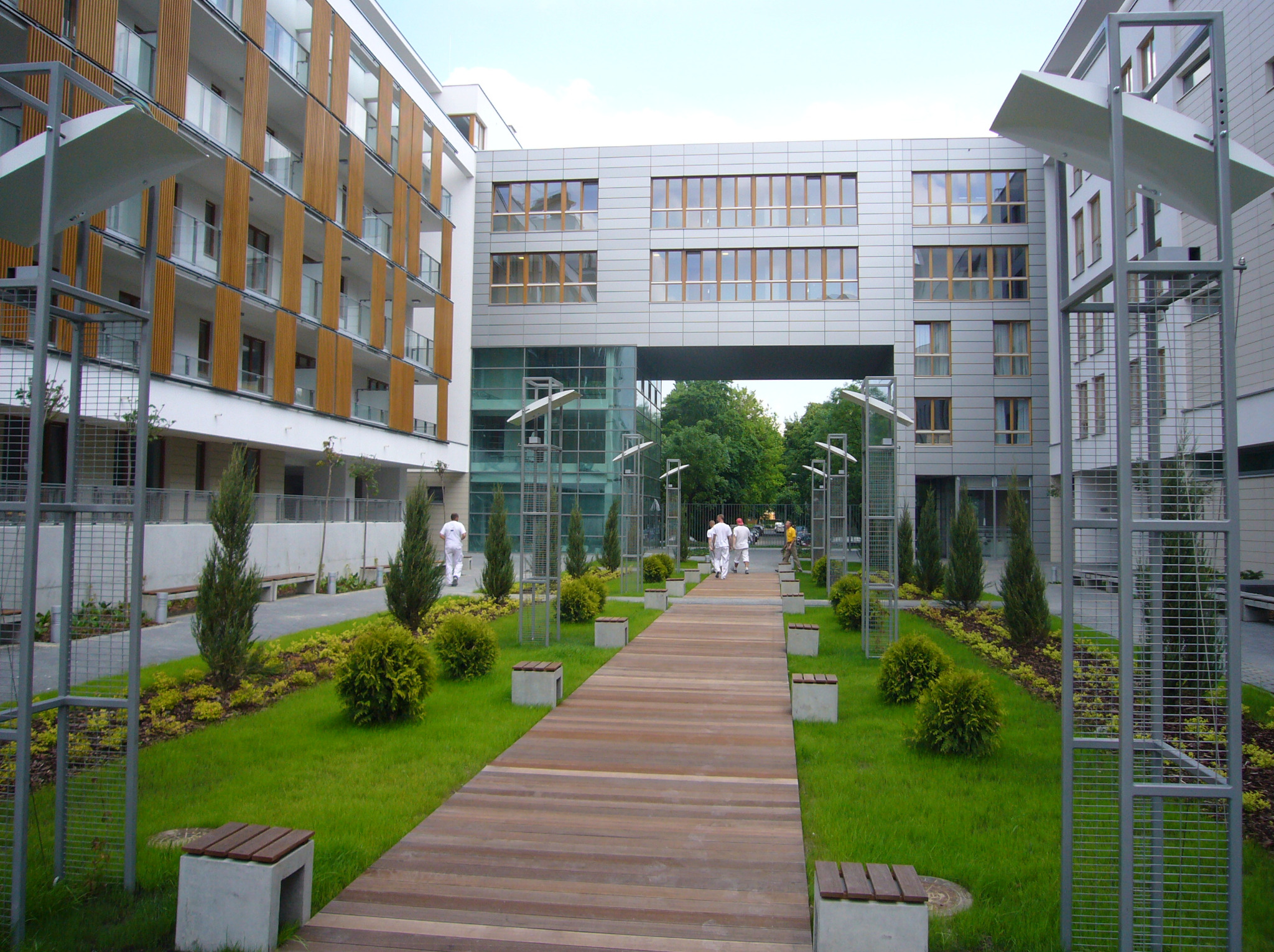 Nowoczesny pasaż między budynkami z drewnianą ścieżką, zielenią i metalowymi konstrukcjami. Widoczne osoby w oddali, jasne oświetlenie dzienne.