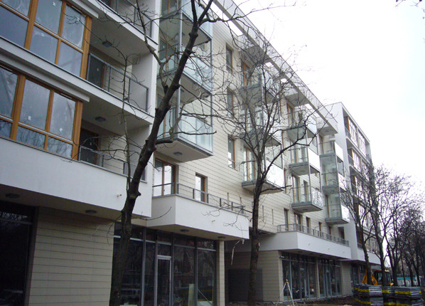 Nowoczesny budynek apartamentowy z balkonami, widok z dołu, drzewa na pierwszym planie, jasna elewacja, duże okna, szklane balustrady balkonów.