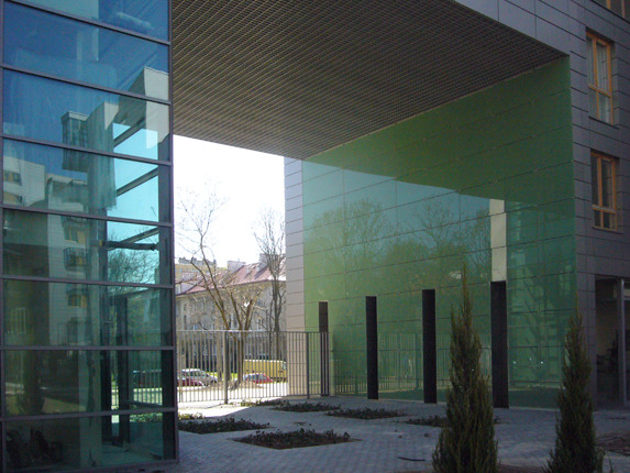 Nowoczesny budynek biurowy z przeszkloną fasadą w odcieniach zieleni i błękitu, widok z zewnątrz na dziedziniec z elementami małej architektury.