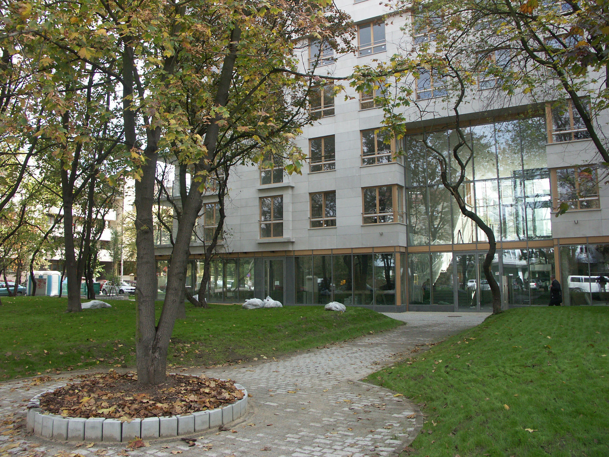 Elewacja budynku biurowego z dużymi oknami, widok z parku z drzewami w jesiennej szacie. Kamienna ścieżka prowadzi do wejścia. Na trawniku leżą białe worki.