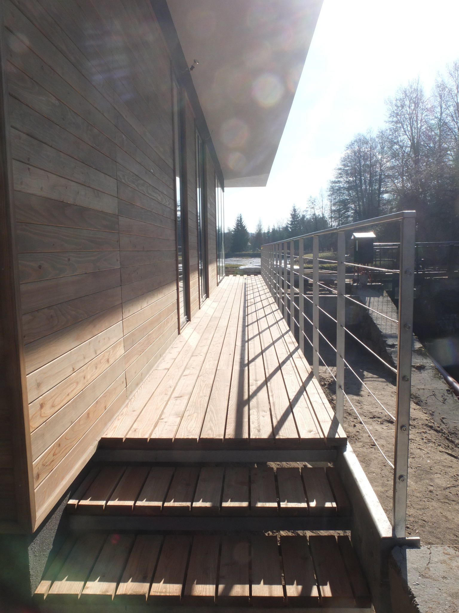 Drewniany taras z metalową balustradą przy budynku z drewnianą elewacją. Widok na zewnątrz, w tle drzewa i woda. Słoneczny dzień.