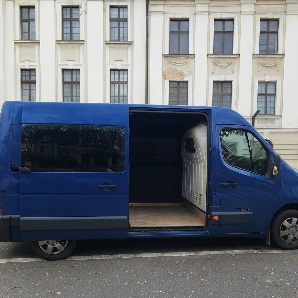 Niebieski bus z otwartymi drzwiami na tle budynku z oknami. Widok z boku, pusta przestrzeń ładunkowa. Gotowy do transportu.