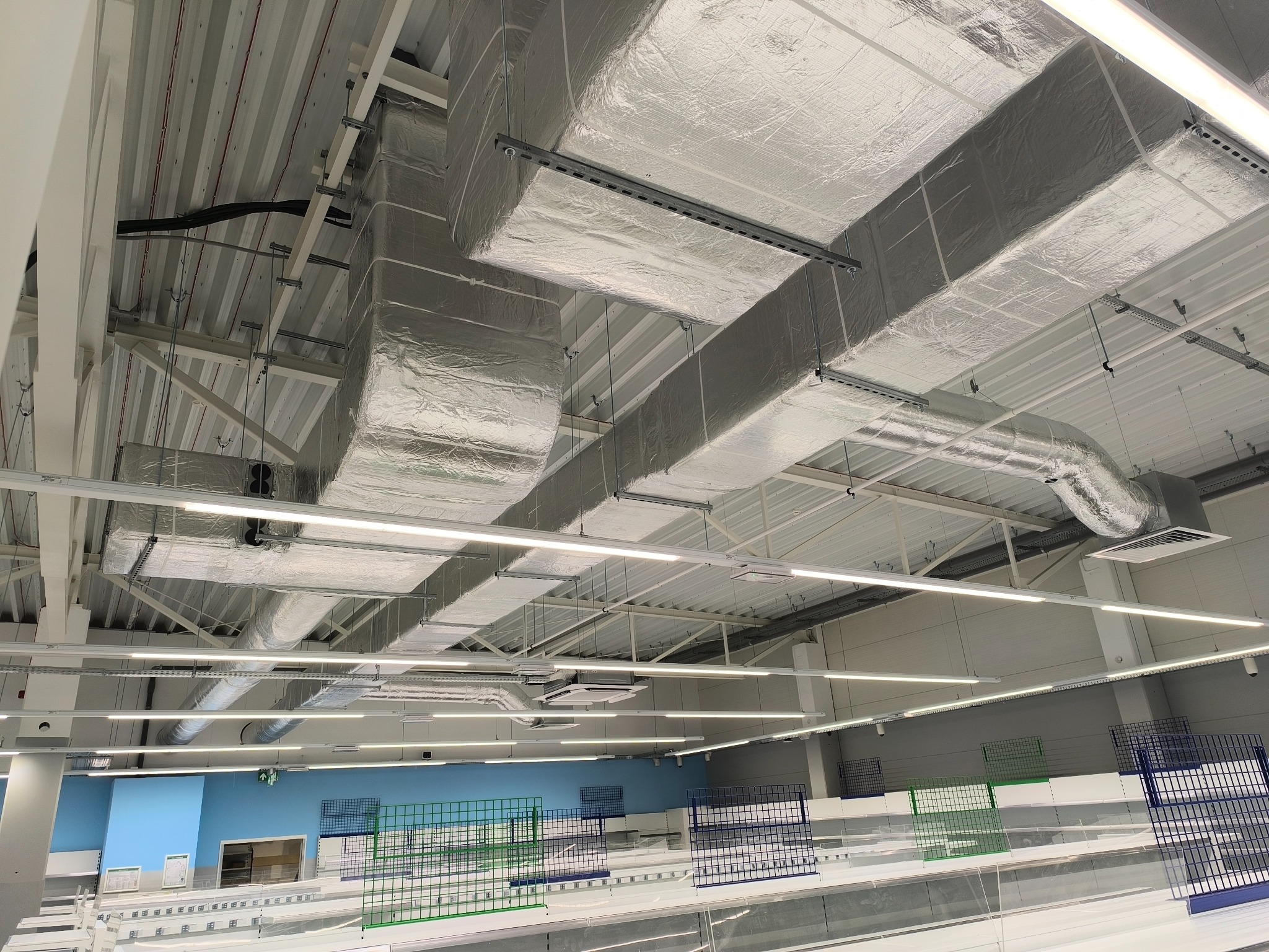 Srebrne, izolowane kanały wentylacyjne pod sufitem hali, z widocznymi elementami mocującymi. W tle regały sklepowe i oświetlenie liniowe.