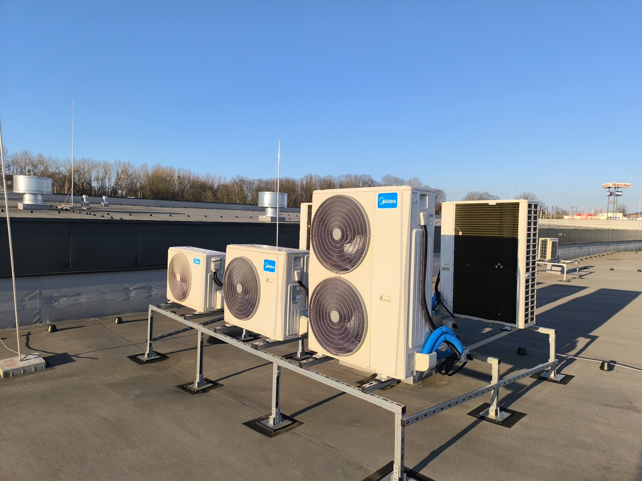 Instalacja klimatyzacji Midea na dachu budynku. Widoczne jednostki zewnętrzne na metalowej konstrukcji wsporczej, błękitne niebo w tle. Ujęcie z niskiego kąta.