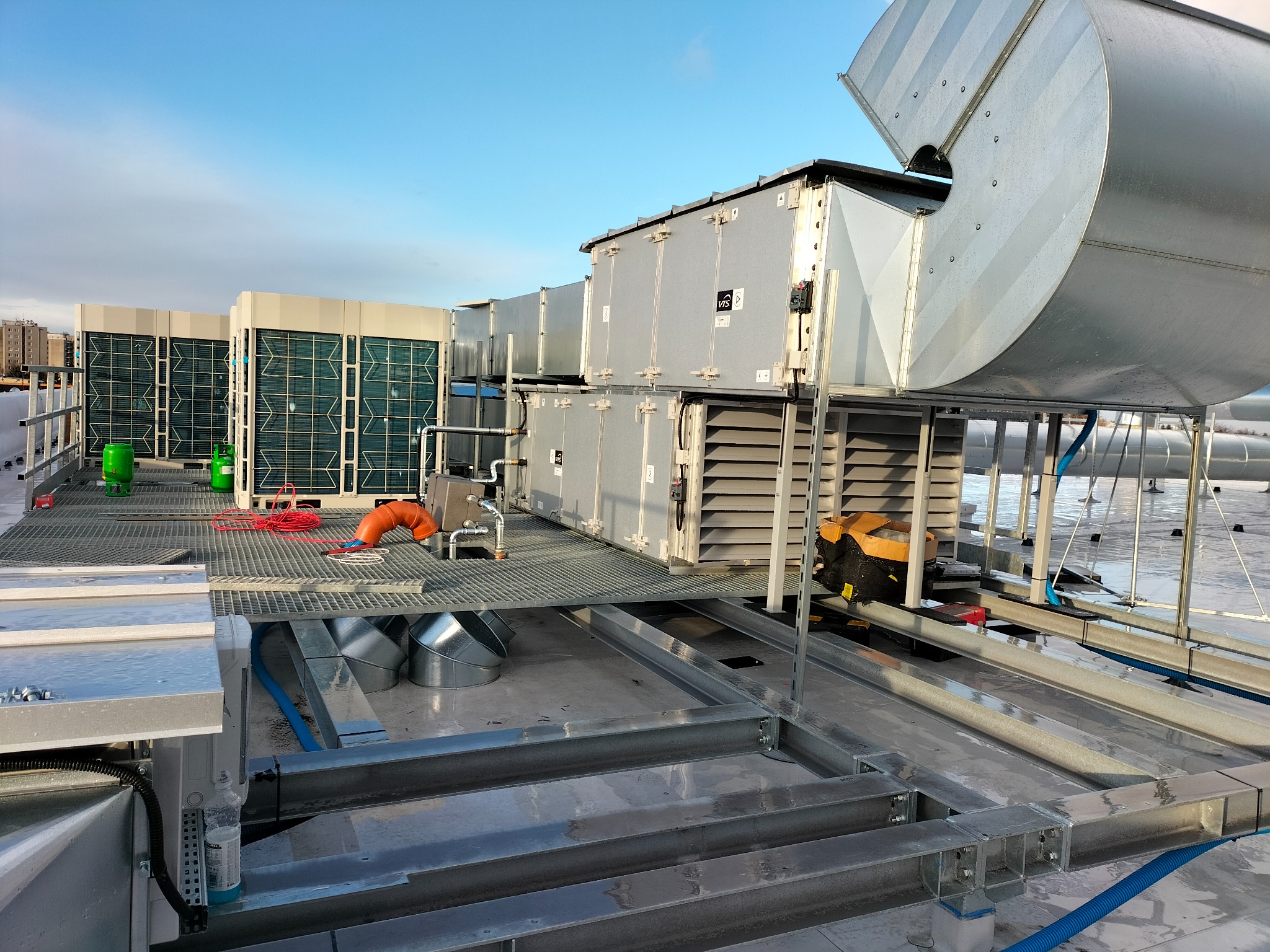 Instalacja wentylacji na dachu budynku z widocznymi elementami systemu, w tym kanałami wentylacyjnymi i klimatyzatorami. Widok z góry, metalowa konstrukcja.