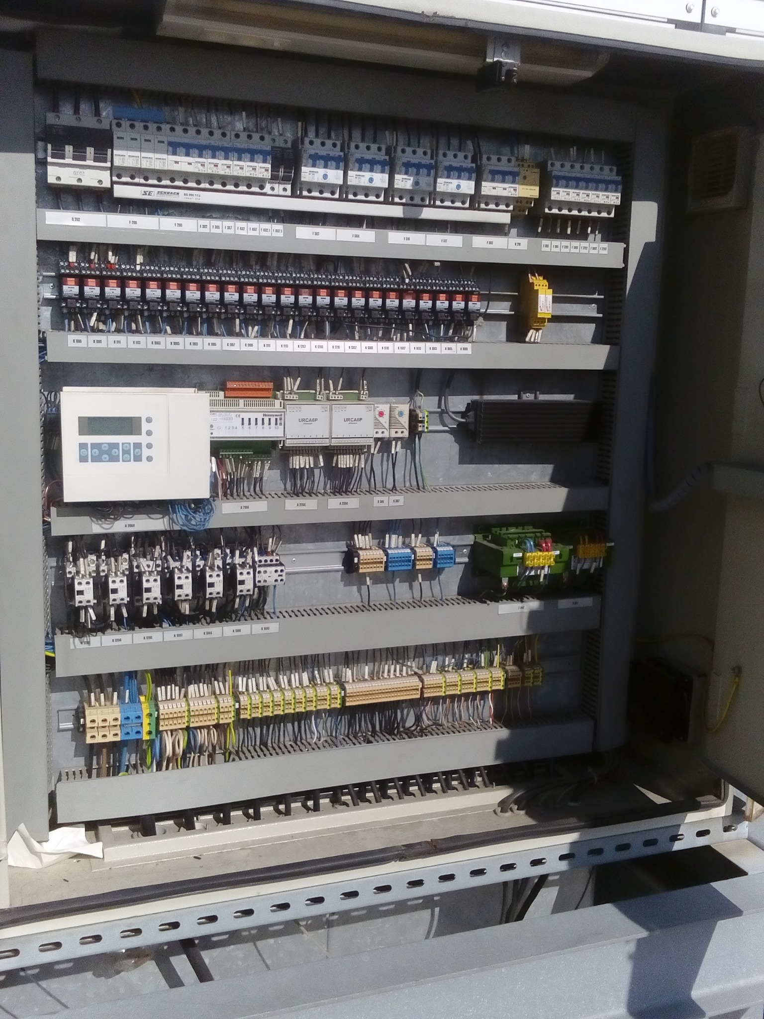 Otwarta szafa sterownicza z licznymi bezpiecznikami, przekaźnikami i przewodami. Widoczne elementy automatyki i sterowania. Szara obudowa, uporządkowany układ komponentów.