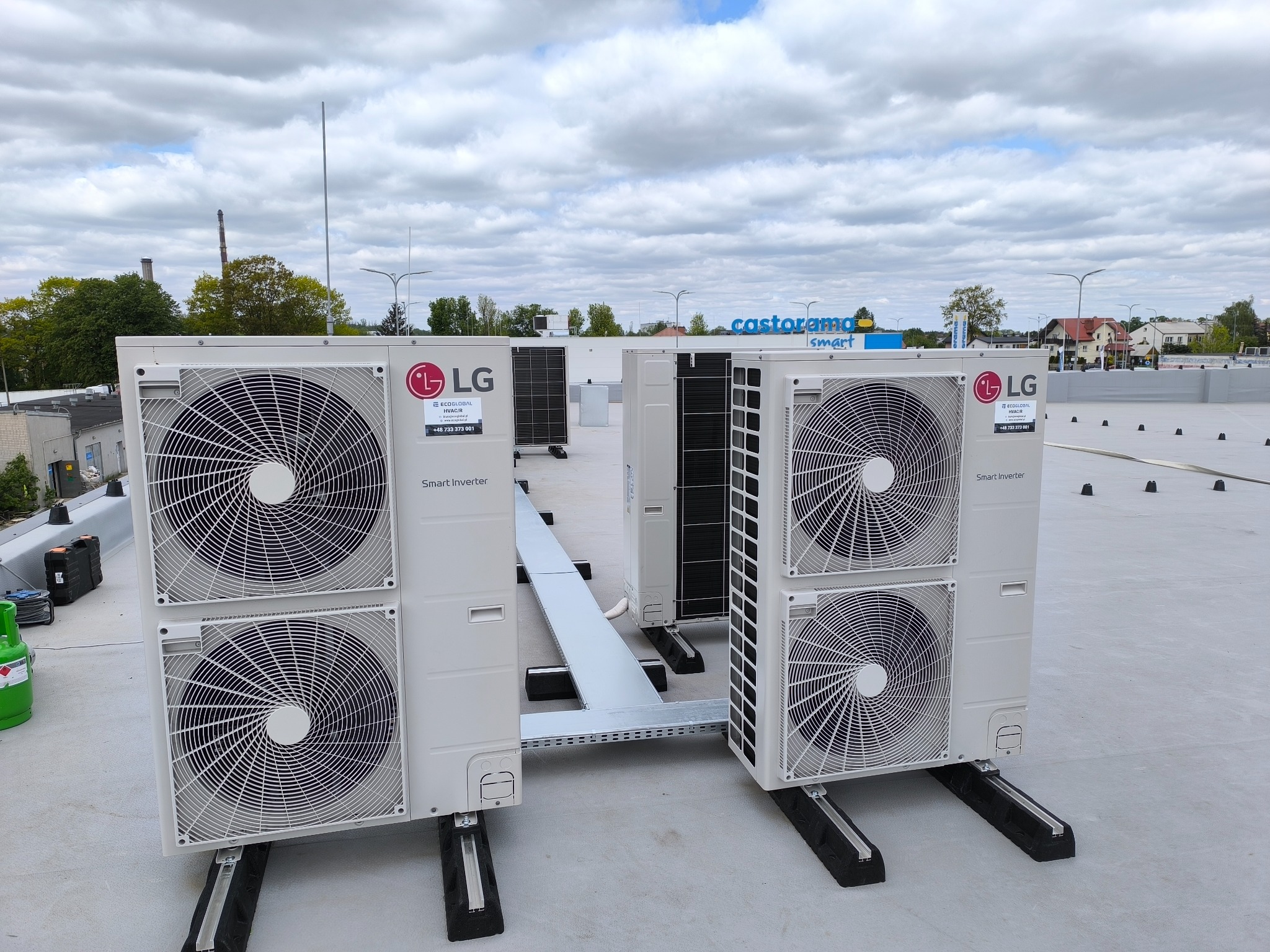 Dwa zestawy klimatyzatorów LG Smart Inverter na dachu budynku, połączone kanałem. W tle widoczny budynek Castorama Smart i pochmurne niebo.