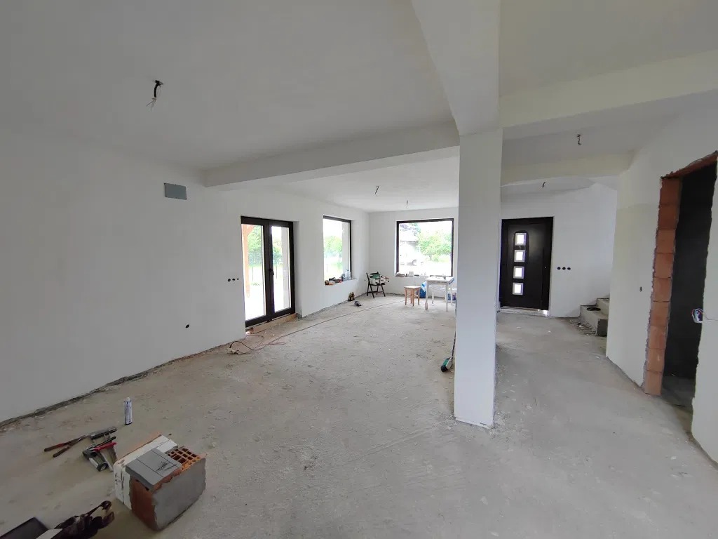 Wnętrze domu w stanie deweloperskim w Jabłonnie Lackiej. Białe ściany, surowa podłoga, widoczne narzędzia i materiały budowlane. Okna i drzwi zamontowane.