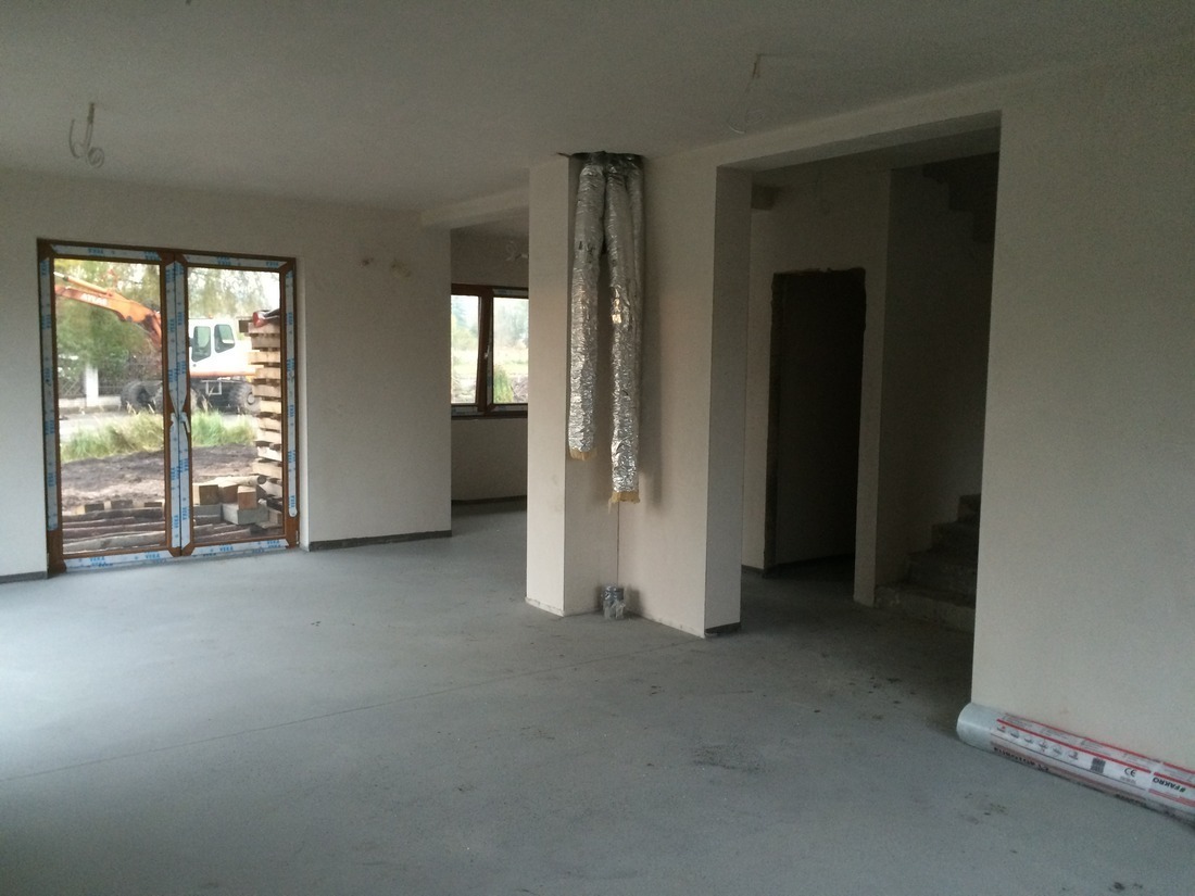 Surowy stan wnętrza domu w trakcie budowy: widoczne okna, instalacje wentylacyjne i wylewka podłogowa. Na zewnątrz koparka.