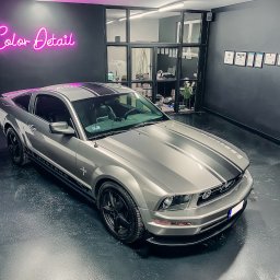 COLOR DETAIL SPÓŁKA Z OGRANICZONĄ ODPOWIEDZIALNOŚCIĄ - Szary Ford Mustang z czarnymi pasami w studiu detailingowym, pod neonem 'Color Detail'. W tle przeszklone biuro i certyfikaty na ścianie. Błyszcząca podłoga.