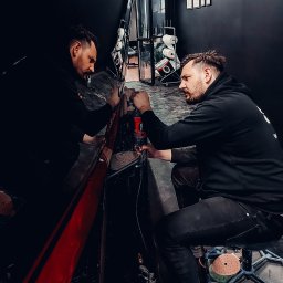 COLOR DETAIL SPÓŁKA Z OGRANICZONĄ ODPOWIEDZIALNOŚCIĄ - Pracownik w czarnej bluzie poleruje czerwony element karoserii samochodu w warsztacie. Widoczne narzędzia i odbicie w lakierowanej powierzchni.