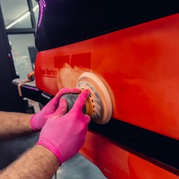 COLOR DETAIL SPÓŁKA Z OGRANICZONĄ ODPOWIEDZIALNOŚCIĄ - Polerowanie czerwonej karoserii samochodu z użyciem polerki rotacyjnej. Ręce w różowych rękawicach. Widoczne ślady pasty polerskiej. Oświetlenie neonowe w tle.