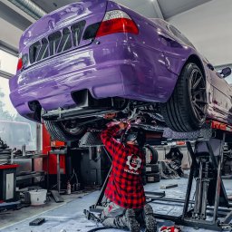 COLOR DETAIL SPÓŁKA Z OGRANICZONĄ ODPOWIEDZIALNOŚCIĄ - Fioletowe auto na podnośniku w warsztacie. Mechanik w czerwonej koszuli spawa podwozie. Widoczne narzędzia i elementy wyposażenia warsztatu.