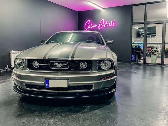 Szary Ford Mustang z czarnymi pasami na masce, zaparkowany w nowoczesnym garażu z neonem 'Color Detail' i przeszklonymi drzwiami.