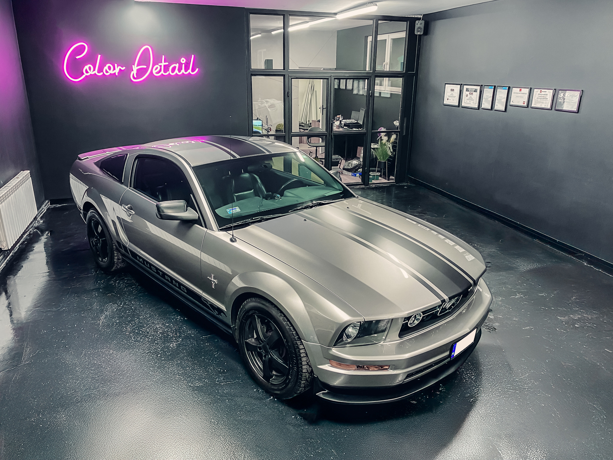 Szary Ford Mustang z czarnymi pasami w studiu detailingowym, pod neonem 'Color Detail'. W tle przeszklone biuro i certyfikaty na ścianie. Błyszcząca podłoga.