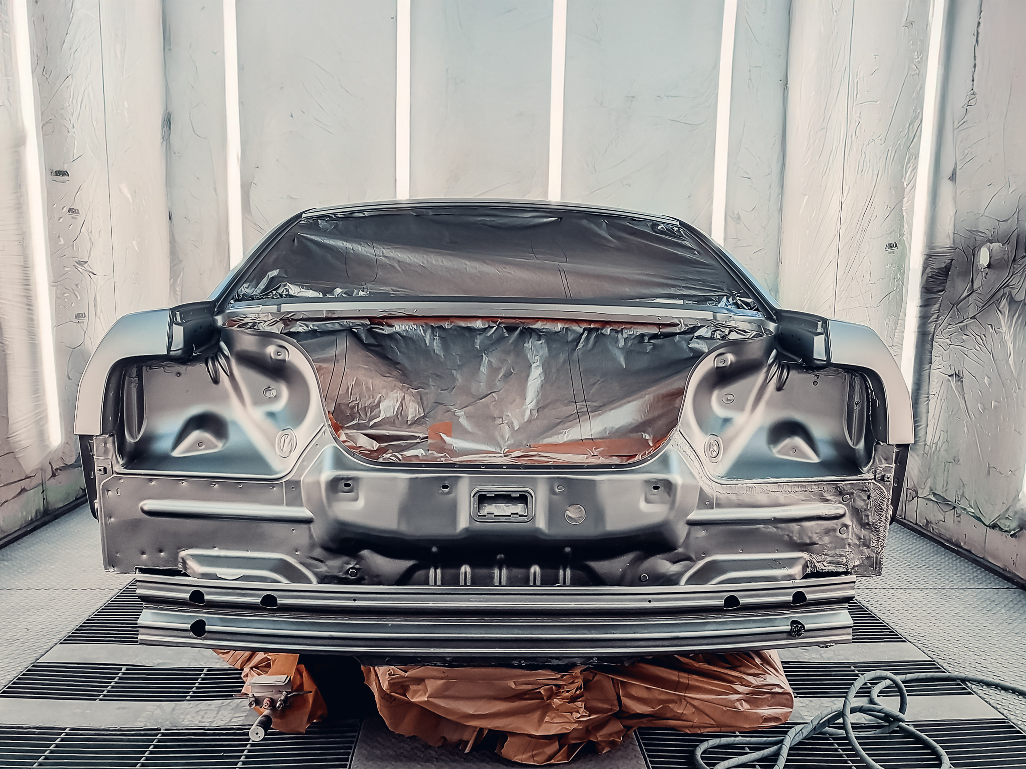 Karoseria auta po piaskowaniu, przygotowana do lakierowania w kabinie lakierniczej. Elementy zabezpieczone folią, widoczny pistolet lakierniczy i przewód.