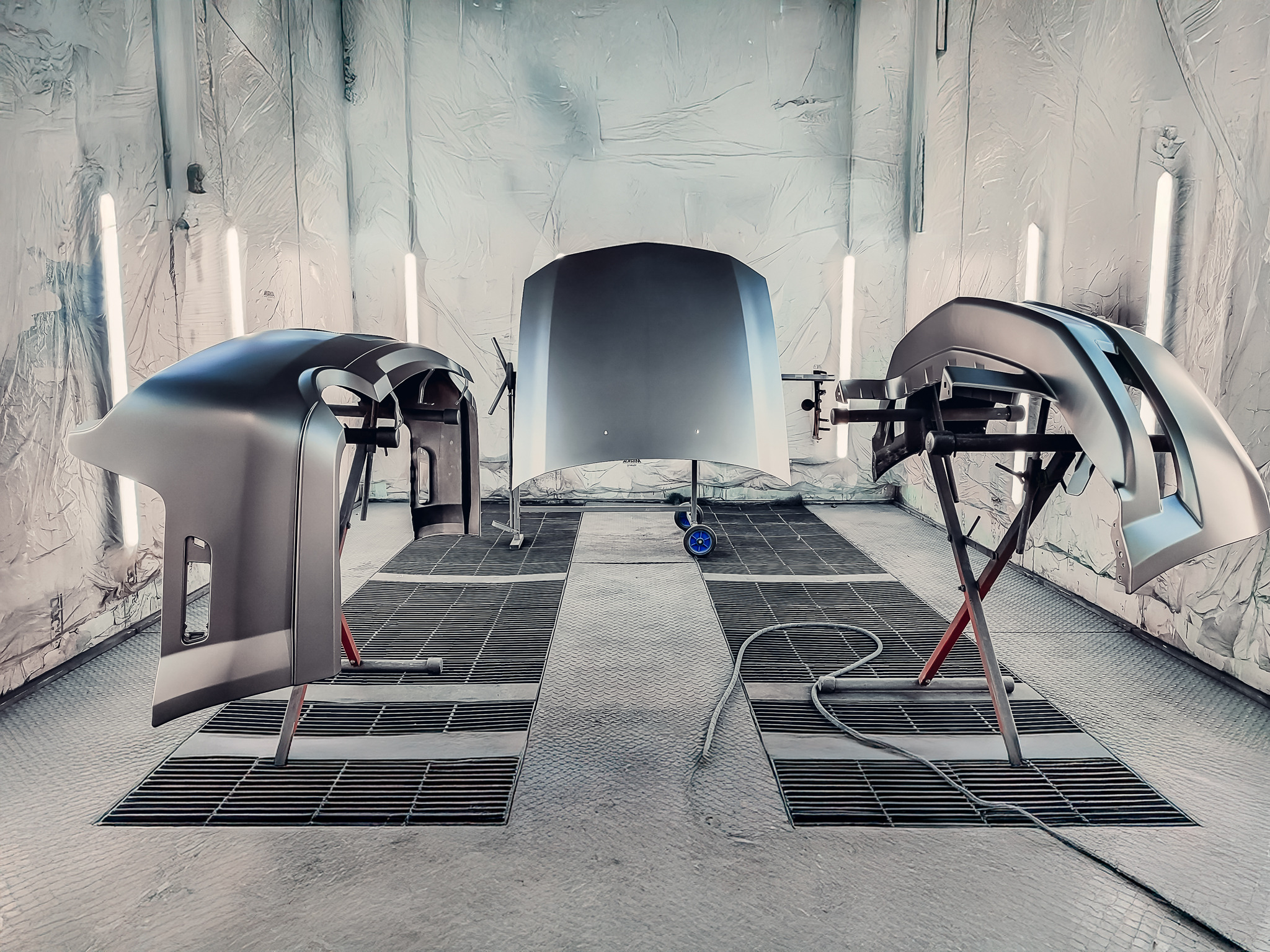 Elementy karoserii (maska, zderzaki) po lakierowaniu w kabinie lakierniczej. Widoczne kraty podłogowe i oświetlenie. Metaliczne wykończenie.