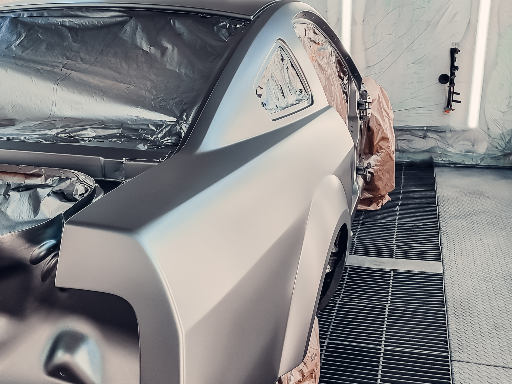 Auto w komorze lakierniczej, przygotowane do malowania. Elementy zabezpieczone folią i papierem. Widoczny fragment karoserii w matowym, szarym kolorze.