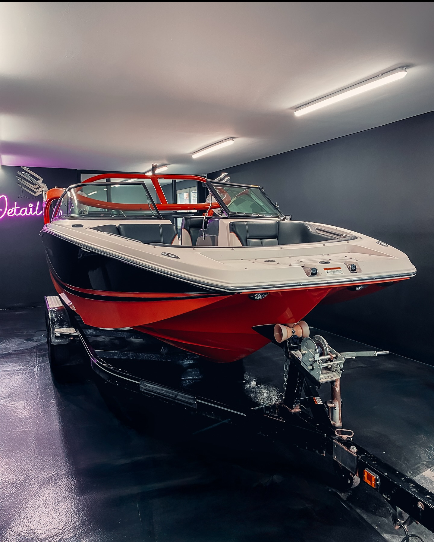 Lśniąca, czerwono-biała łódź motorowa na czarnej przyczepie w sterylnym garażu z neonowym napisem 'Detail'. Perfekcyjny połysk i dbałość o detale.