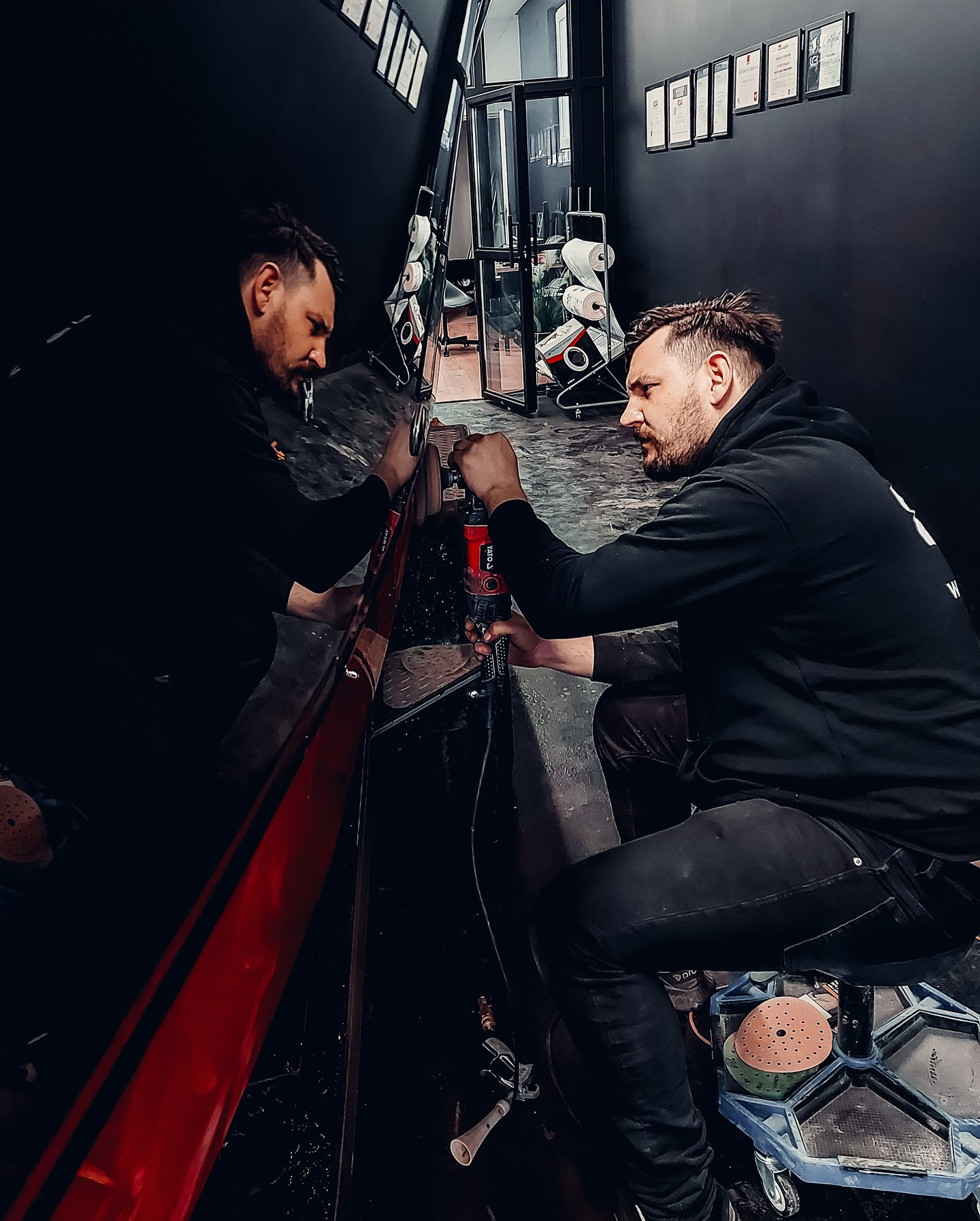 Pracownik w czarnej bluzie poleruje czerwony element karoserii samochodu w warsztacie. Widoczne narzędzia i odbicie w lakierowanej powierzchni.