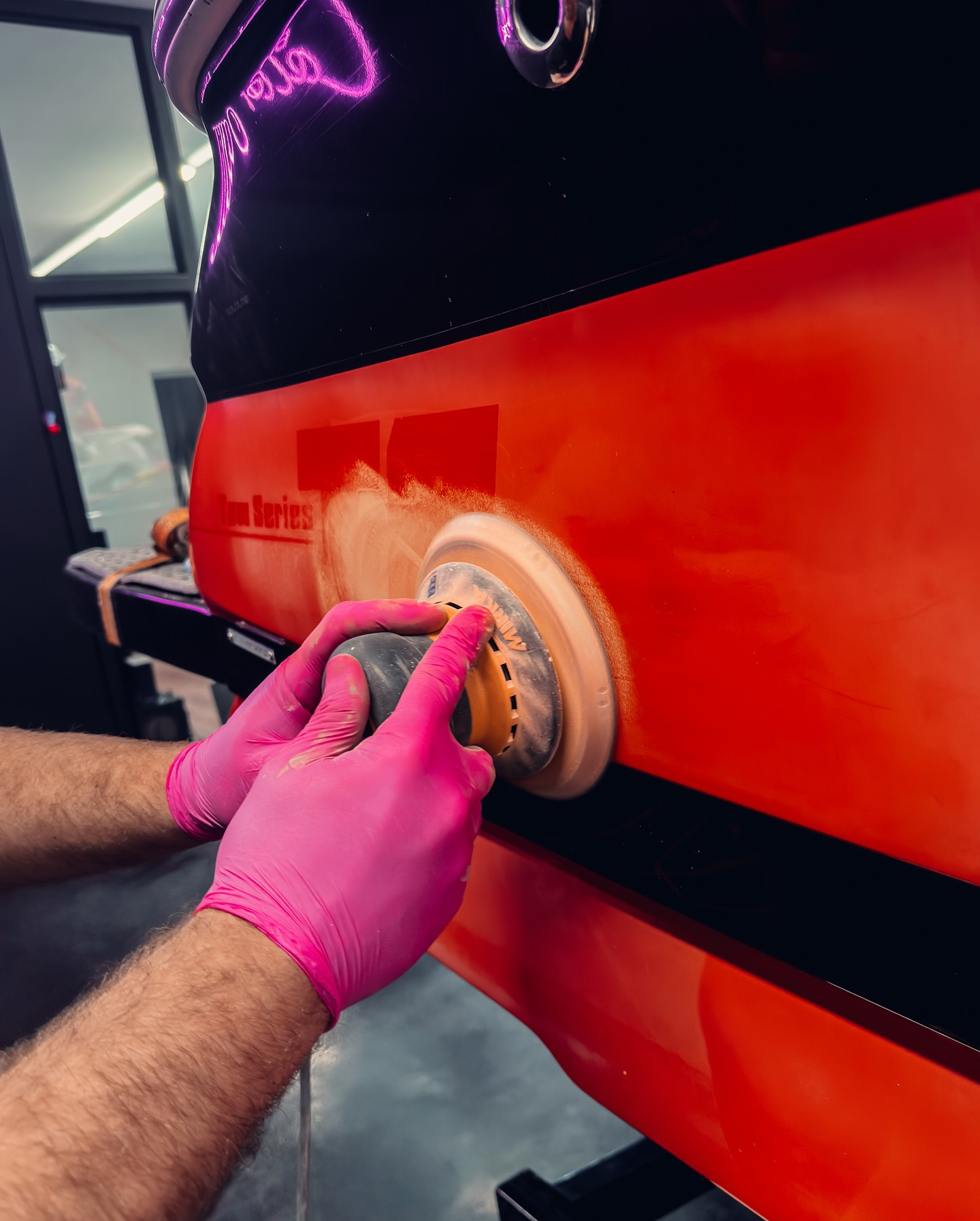 Polerowanie czerwonej karoserii samochodu z użyciem polerki rotacyjnej. Ręce w różowych rękawicach. Widoczne ślady pasty polerskiej. Oświetlenie neonowe w tle.