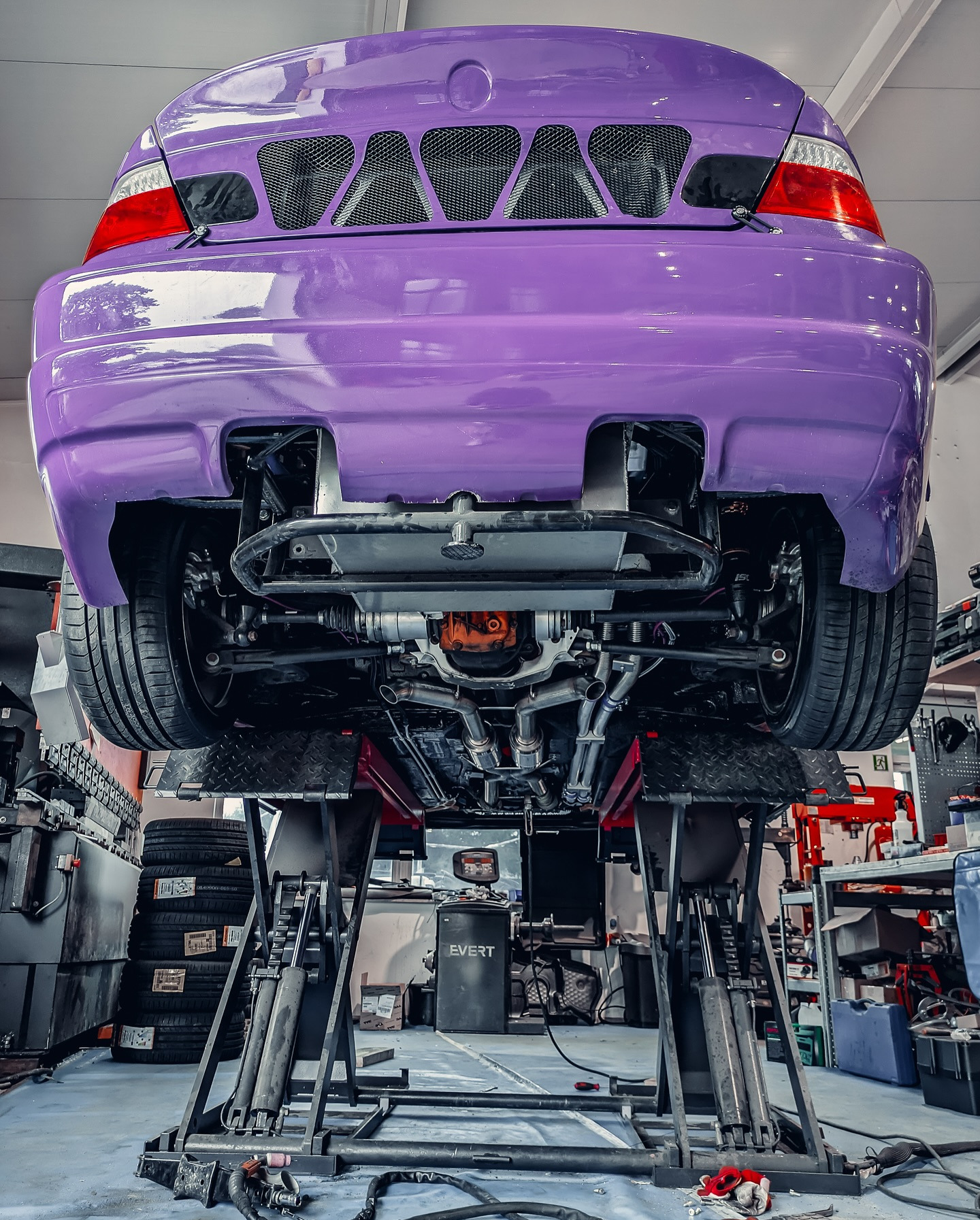 Fioletowy samochód na podnośniku warsztatowym, widok od spodu, z widocznymi elementami zawieszenia i układu wydechowego. Detale tuningu.