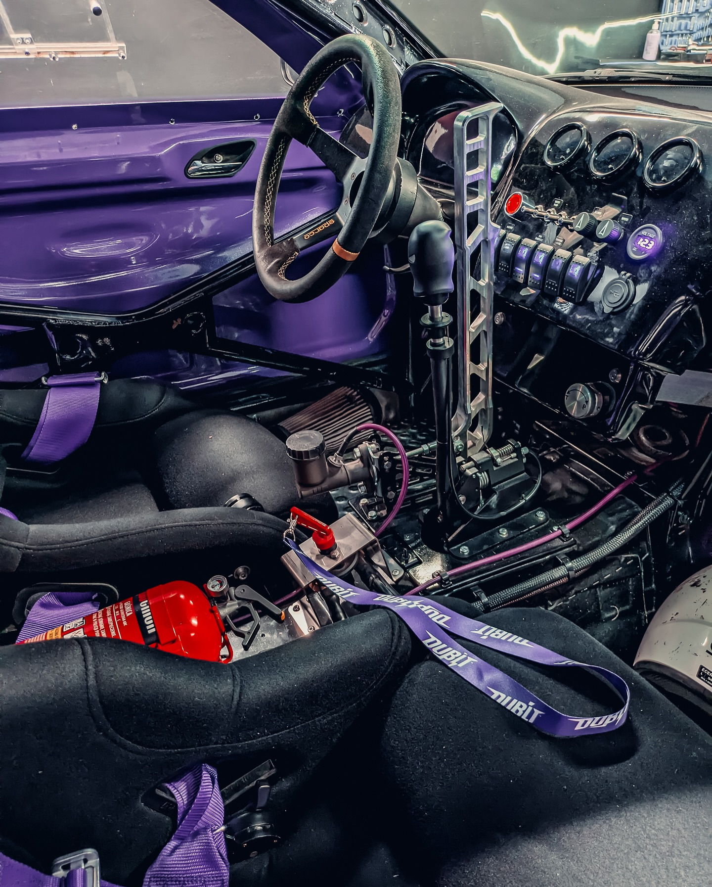 Wnętrze zmodyfikowanego samochodu wyścigowego z fioletowymi akcentami, widoczne fotele kubełkowe, klatka bezpieczeństwa, gaśnica i panel sterowania z przełącznikami.