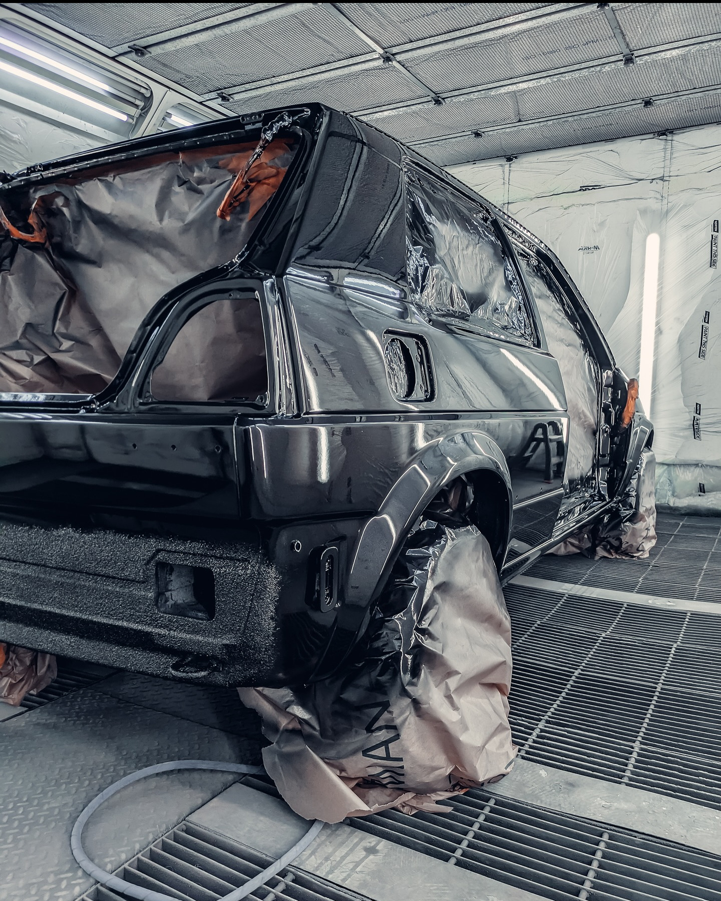 Czarny samochód w kabinie lakierniczej, zabezpieczony folią przed lakierowaniem. Widoczna faktura 'baranka' na dolnej części karoserii. Metalowa podłoga kabiny.