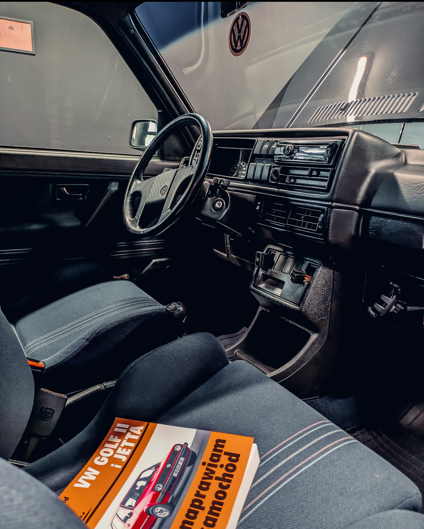 Wnętrze klasycznego VW Golf II: kierownica, deska rozdzielcza z radiem, fotel z książką 'VW Golf II i Jetta - naprawiam samochód'. Widoczna maska i logo VW.