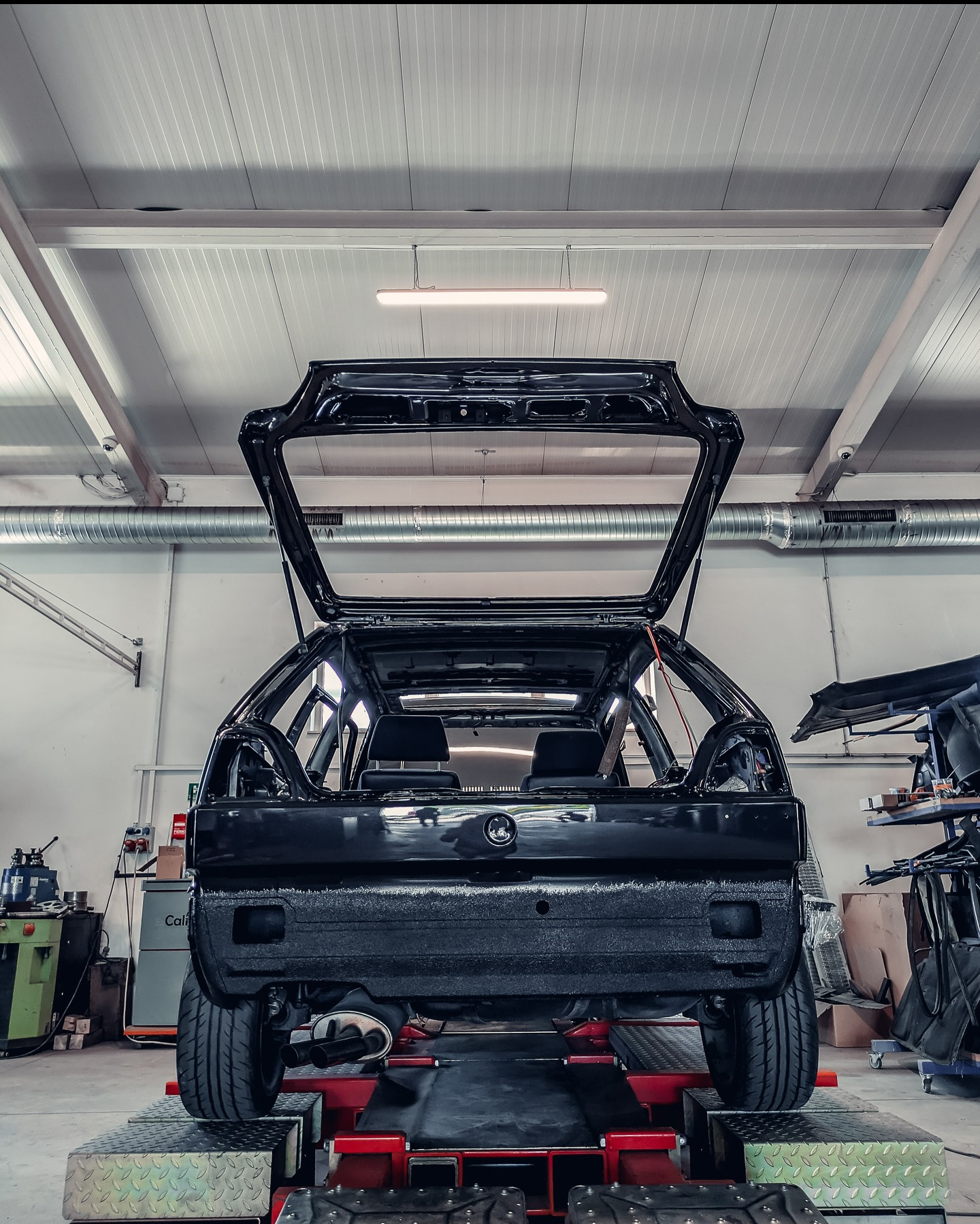 Karoseria czarnego auta na podnośniku w warsztacie. Widoczne elementy wyposażenia warsztatu i oświetlenie. Auto przygotowane do dalszych prac blacharsko-lakierniczych.