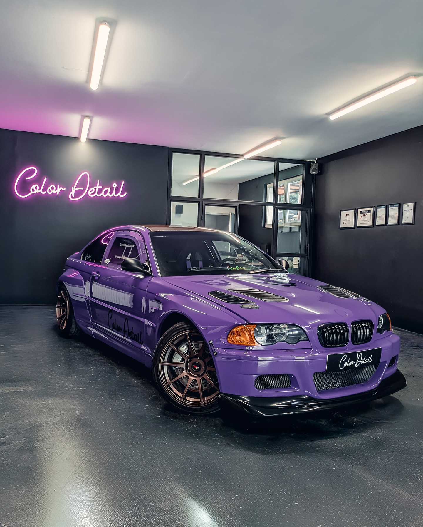 Fioletowe BMW E46 Coupe po tuningu, z brązowymi felgami, w garażu z neonem 'Color Detail' i certyfikatami na ścianie. Unikalny kolor i body kit.