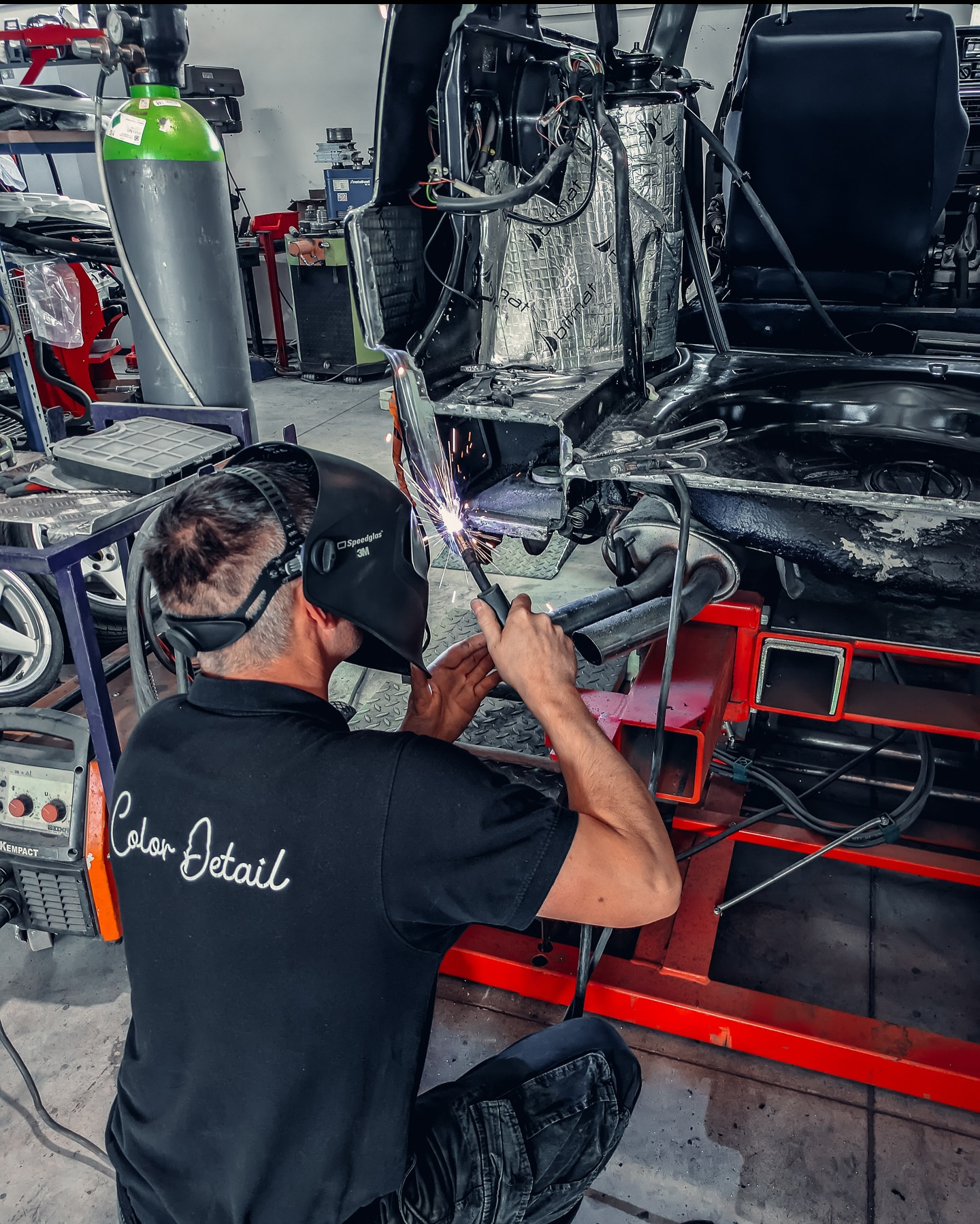 Spawanie karoserii auta w warsztacie. Mężczyzna w masce spawalniczej 3M łączy elementy konstrukcyjne. Widoczny spawacz, spawarka Kempack i butla z gazem.
