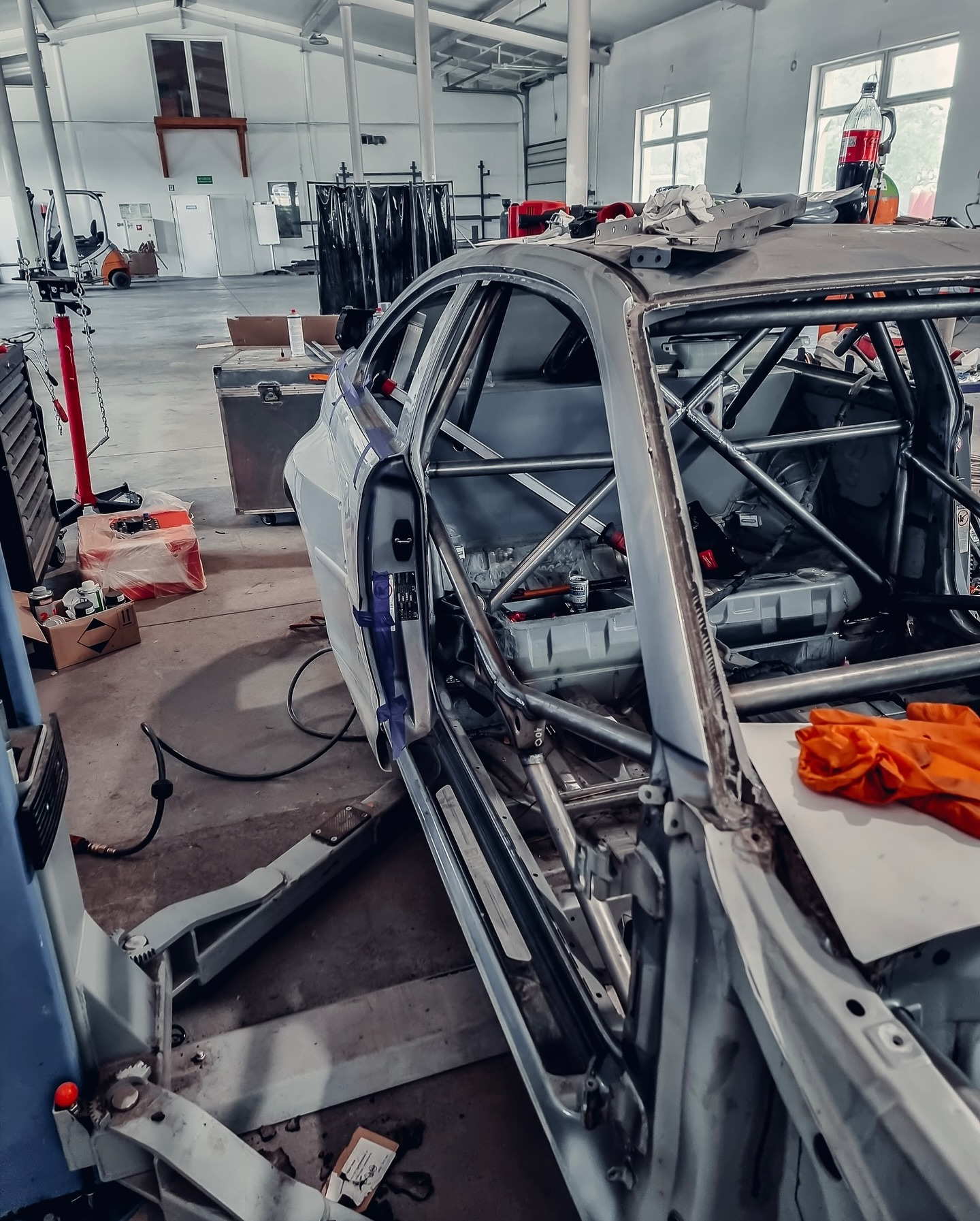 Karoseria samochodu sportowego w trakcie budowy klatki bezpieczeństwa, widoczna na podnośniku w warsztacie. Elementy karoserii zabezpieczone taśmą.