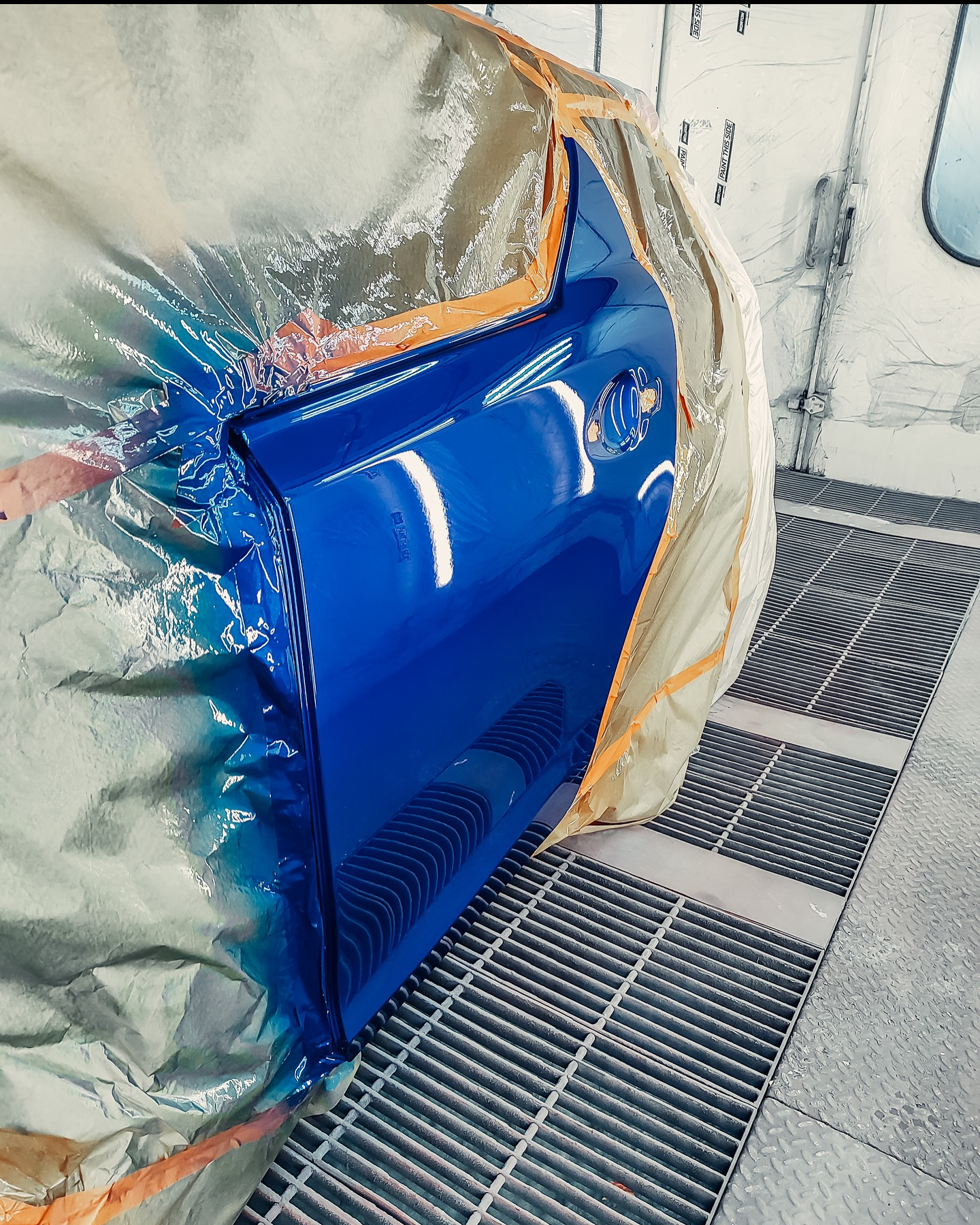 Błyszczące, niebieskie drzwi samochodu po lakierowaniu, częściowo zakryte folią ochronną w kabinie lakierniczej. Widoczna kratka podłogowa.