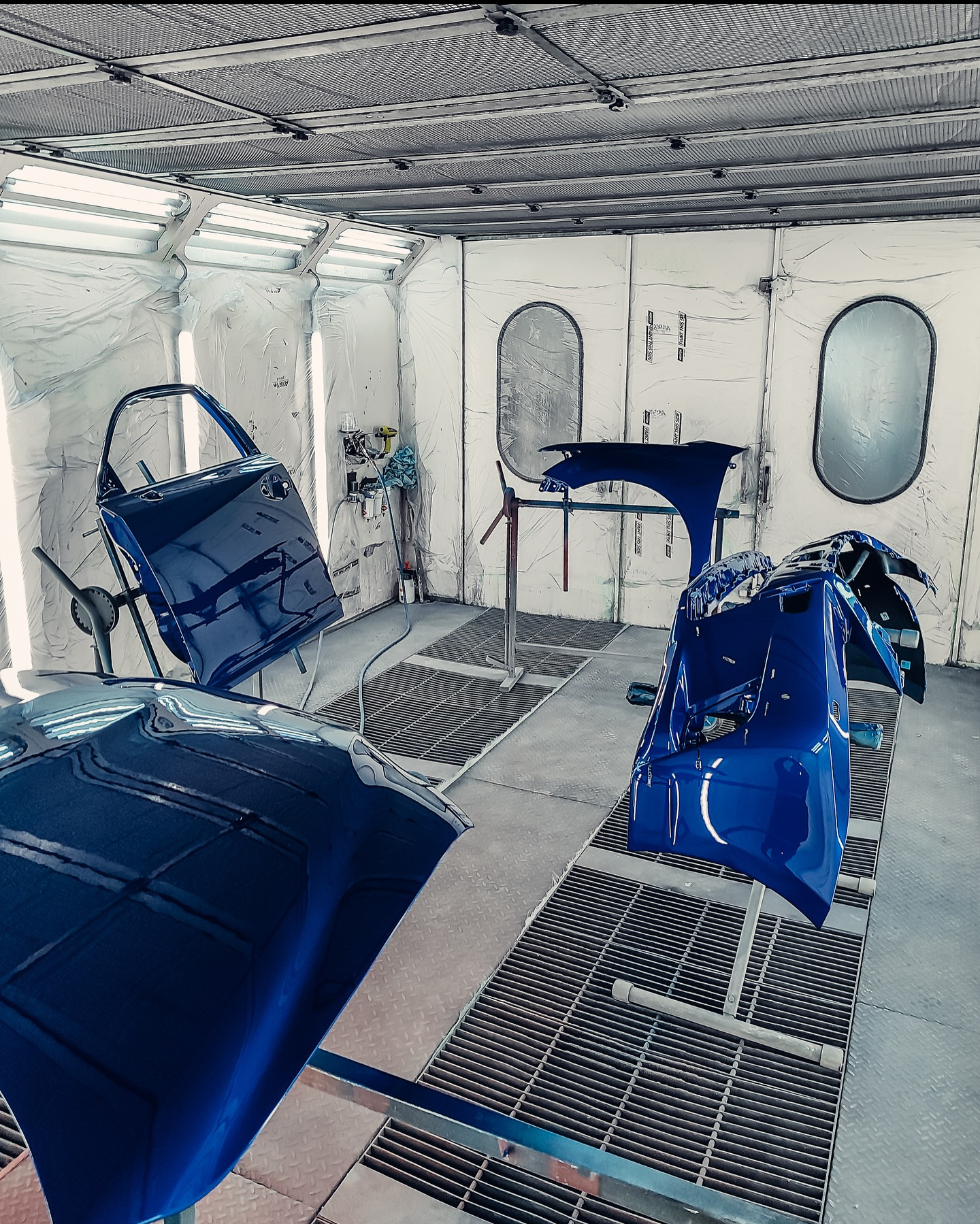Błyszczące, niebieskie elementy karoserii samochodowej (drzwi, błotnik, zderzak) w kabinie lakierniczej, przygotowane do montażu. Widoczne oświetlenie i kratownica podłogowa.