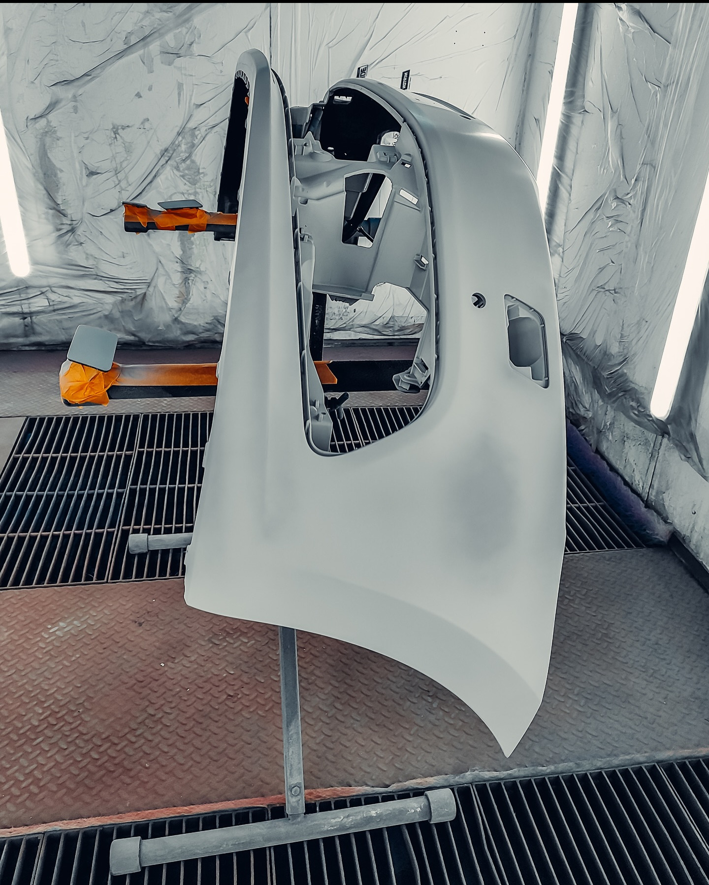 Pomalowany na szaro element karoserii samochodowej, prawdopodobnie błotnik, umieszczony na stojaku w lakierni. Widoczne detale mocowań i otwory technologiczne.