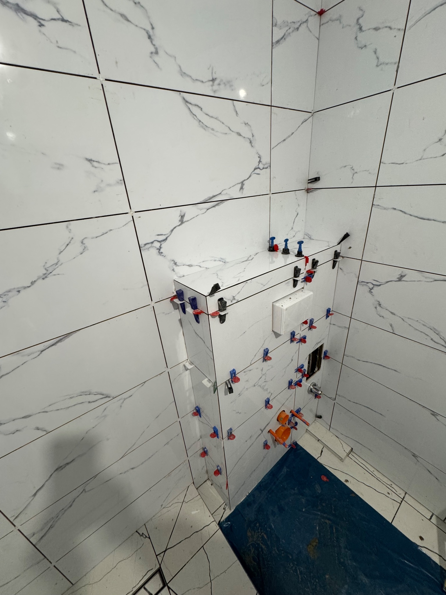 Białe płytki imitujące marmur na ścianach łazienki, z systemem poziomowania płytek. Widoczny fragment podłogi zabezpieczony folią. Ujęcie z góry, z bliska.