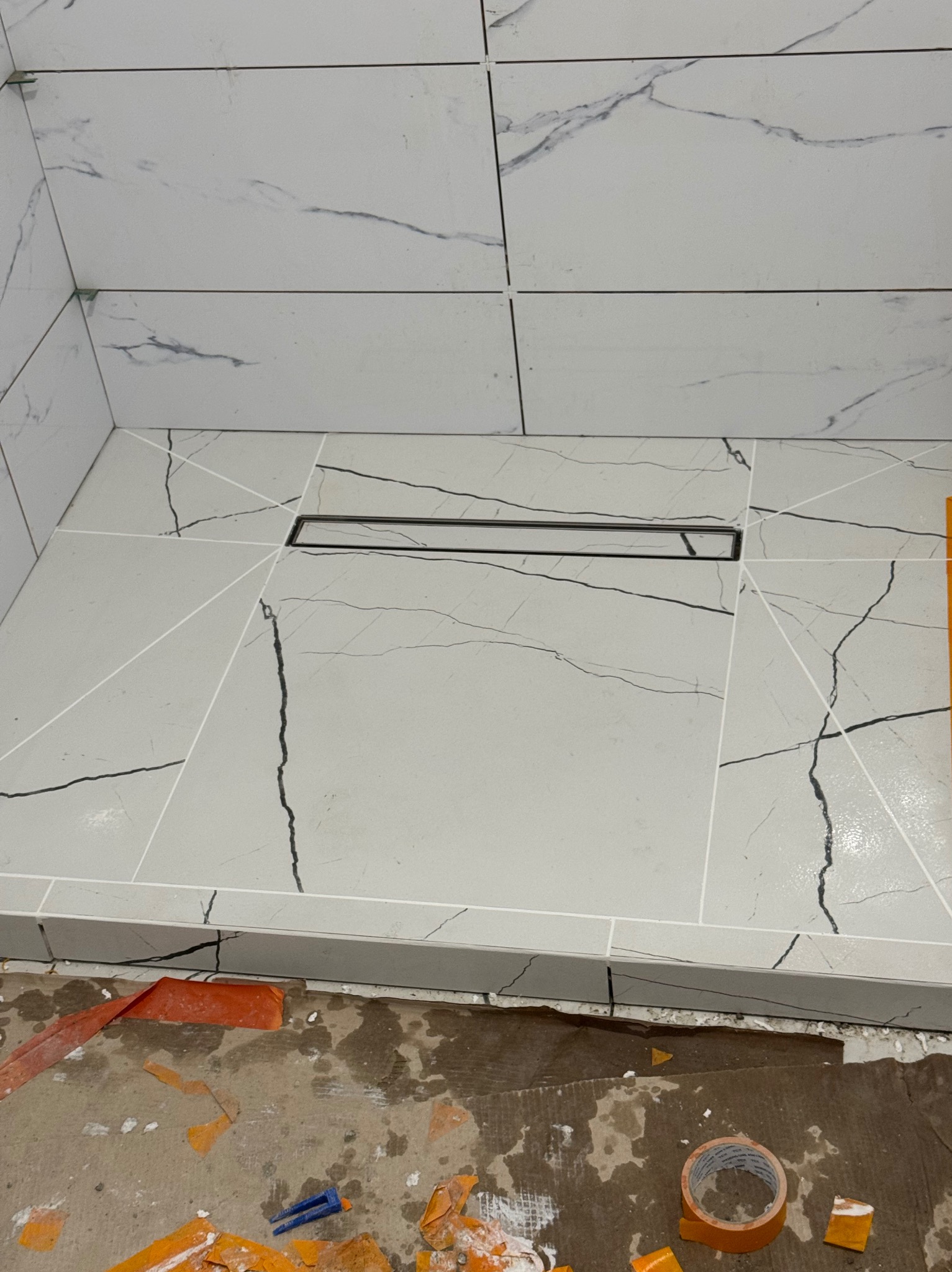 Ukończona strefa prysznicowa z odpływem liniowym, wyłożona płytkami imitującymi marmur z wyraźnym wzorem żyłkowania. Widoczne narzędzia glazurnicze.