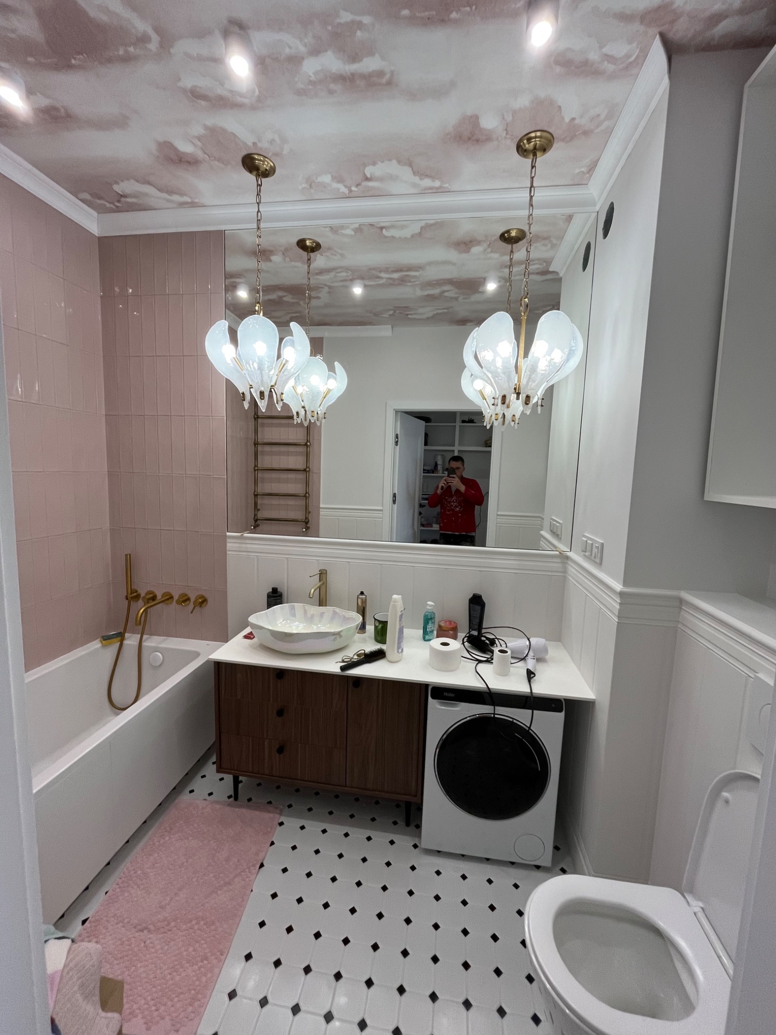 Ukończona łazienka w odcieniach różu i bieli z wzorzystą podłogą. Uwagę przykuwa designerski żyrandol i oryginalny wzór na suficie. W lustrze odbija się osoba robiąca zdjęcie.