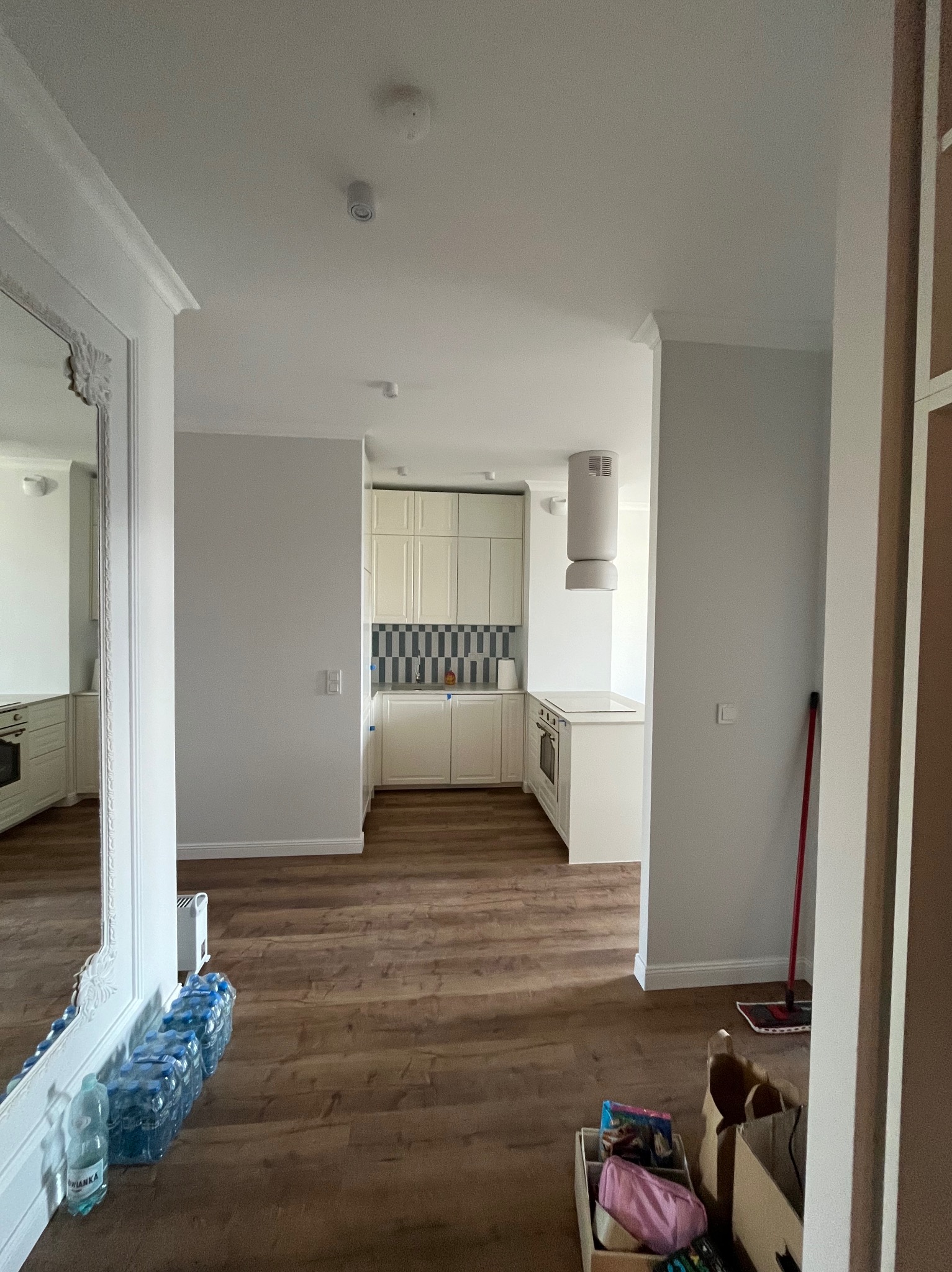 Jasne wnętrze po remoncie: parkiet, białe ściany, kuchnia w tle. Lustro z ozdobną ramą i butelki wody w rogu. Widok na odnowioną przestrzeń.