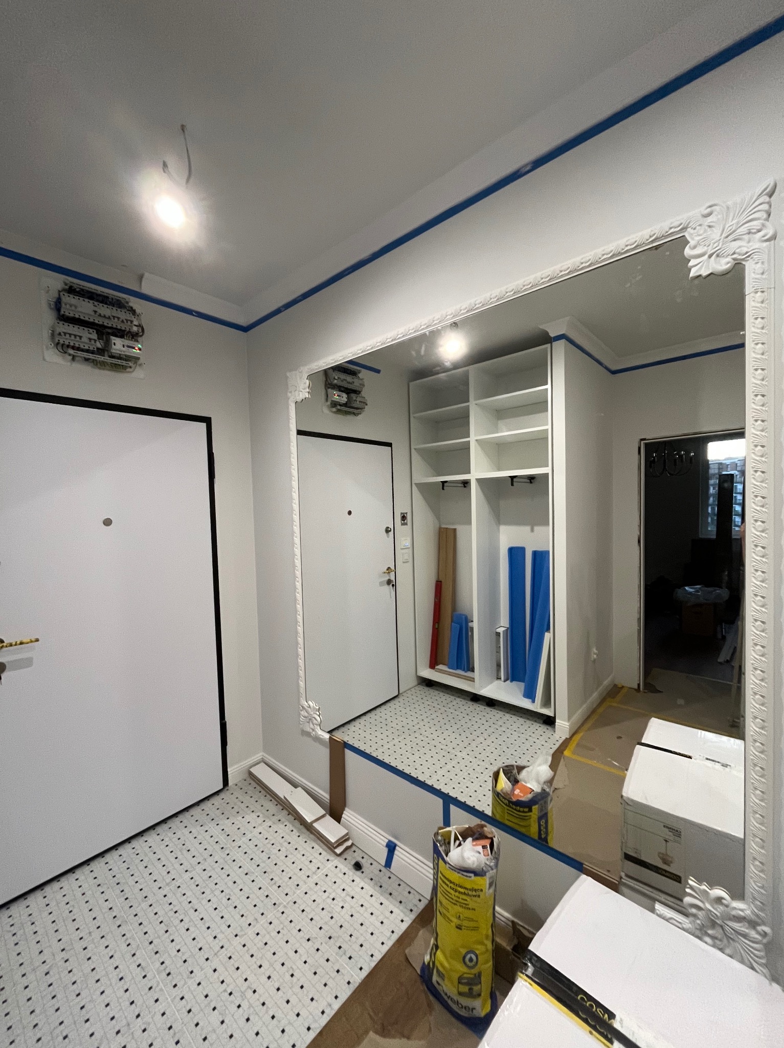 Remont przedpokoju w Warszawie: białe ściany z niebieskim akcentem, lustro w ozdobnej ramie, płytki podłogowe w kropki i szafa wnękowa w trakcie montażu.