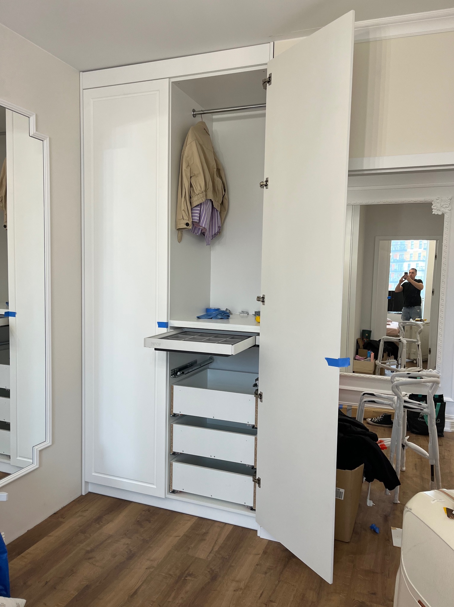 Biała szafa wnękowa z otwartymi drzwiami i wysuwanymi szufladami, widoczna od frontu. W tle lustro z odbijającą się osobą robiącą zdjęcie.
