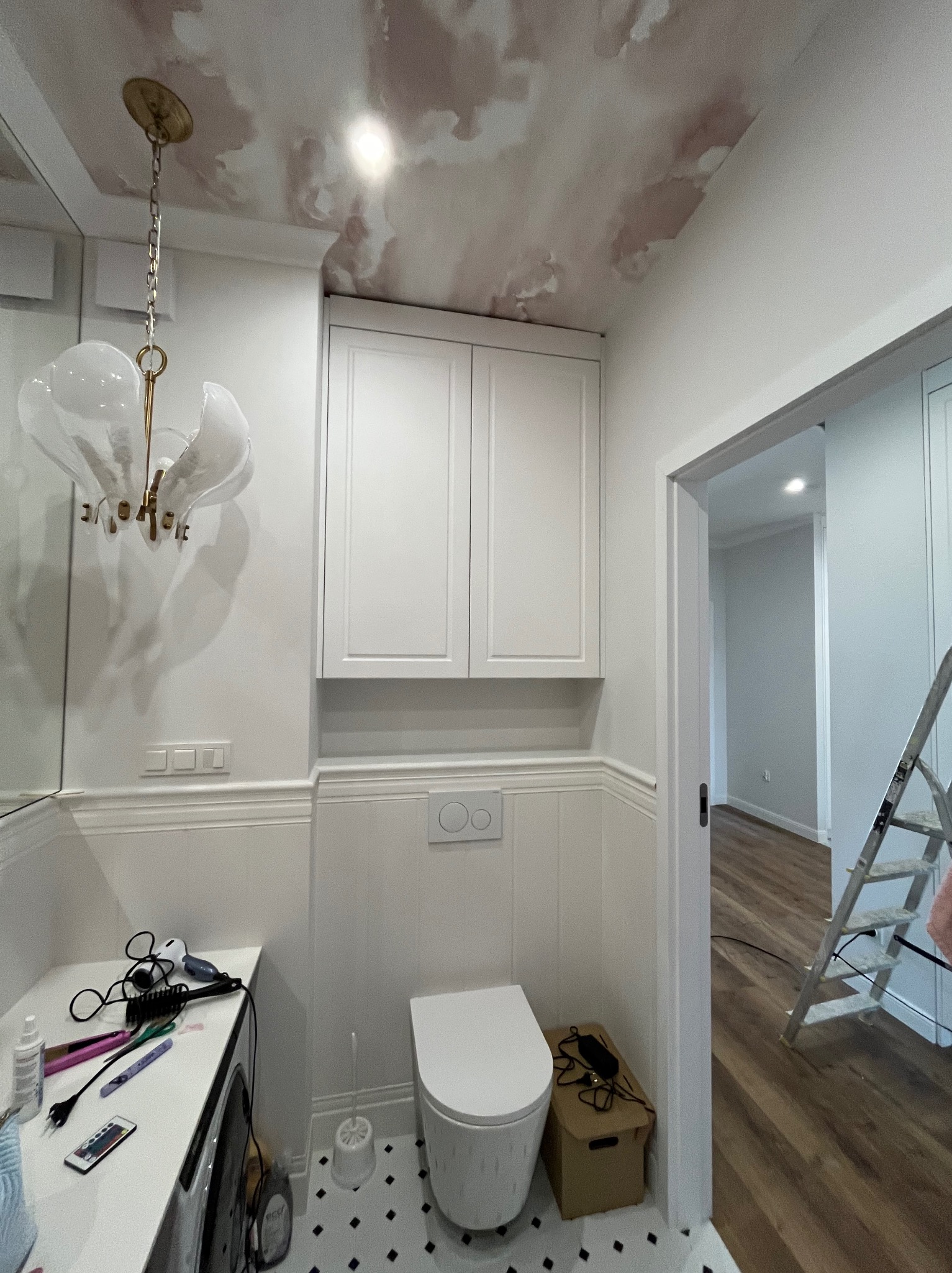 Biała łazienka z wzorzystym sufitem i podłogą w romby. Umywalka z akcesoriami, toaleta i szafka wisząca. W tle widoczna drabina.