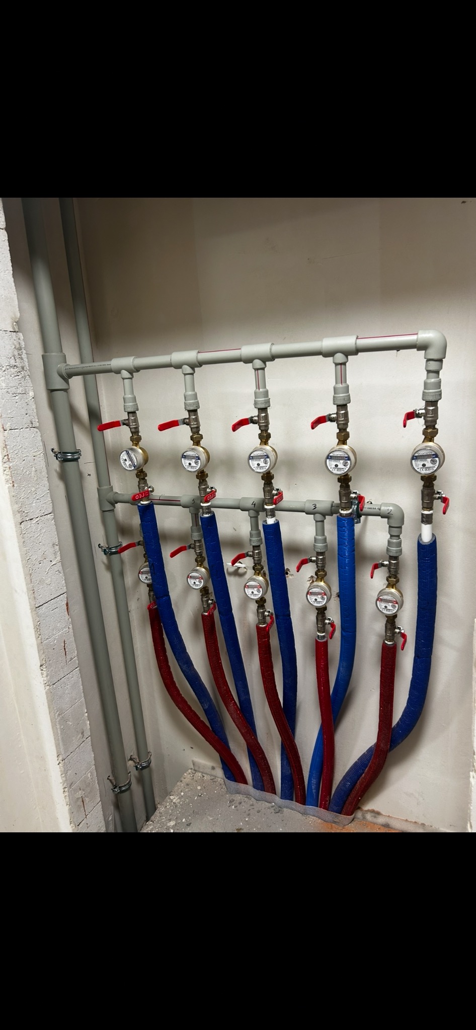 Rozdzielacz instalacji CO i CWU z zaworami, licznikami ciepła i izolowanymi rurami w kolorach niebieskim i czerwonym. Szare rury pionowe przy ścianie.