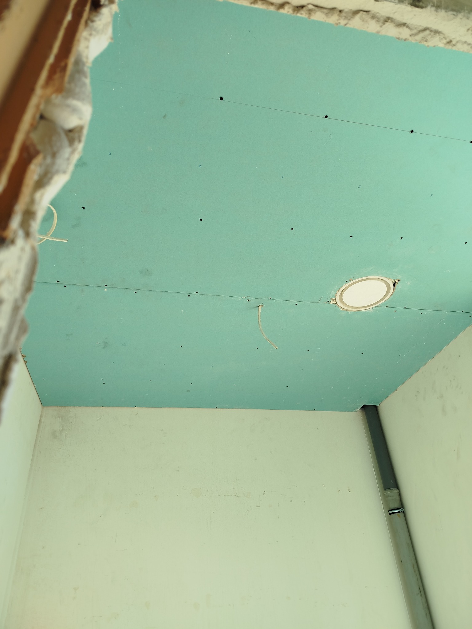 Niebieska płyta karton-gips na suficie z otworem na oświetlenie, widoczne ślady po szpachlowaniu i fragment ściany z rurą. Ujęcie z dołu, w trakcie prac wykończeniowych.