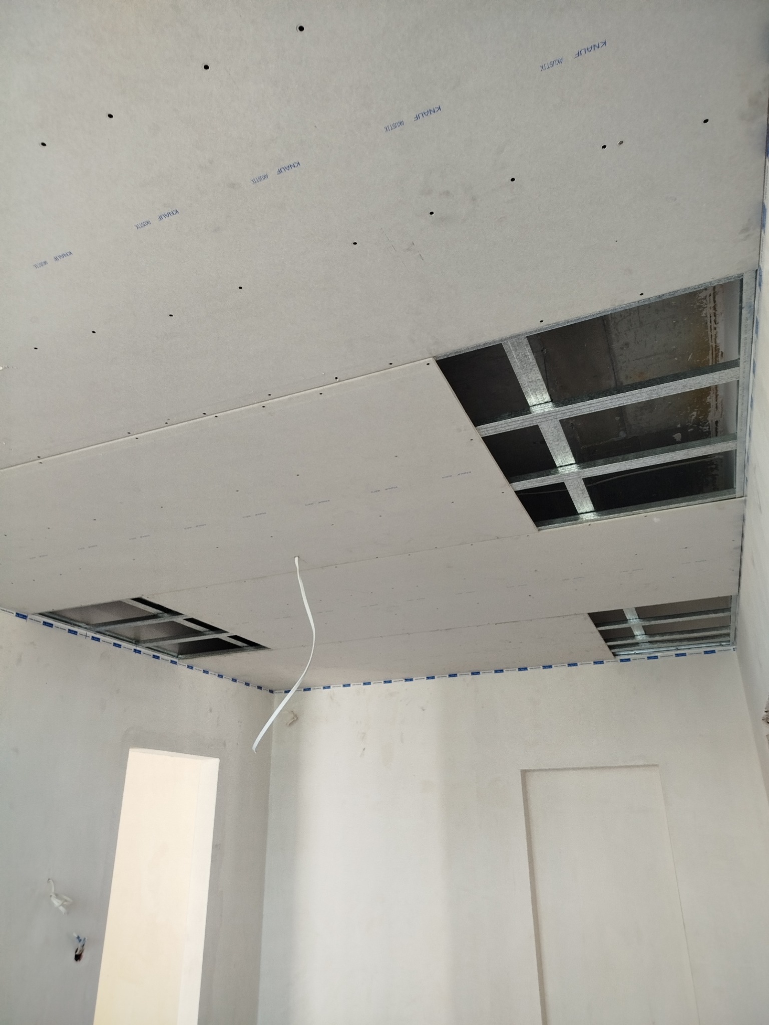 Sufit z płyt gipsowo-kartonowych w trakcie montażu, widoczne metalowe profile i otwory. Na płytach widoczne napisy Knauf Akustik. Białe ściany w tle.